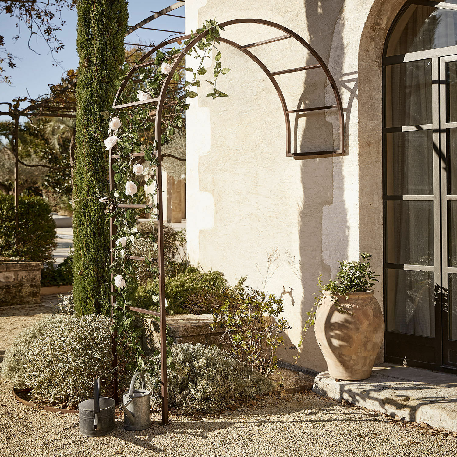 Braune Metall-Gartenpergola mit Bogen, weißen Rosen und Gießkannen auf Kiesboden