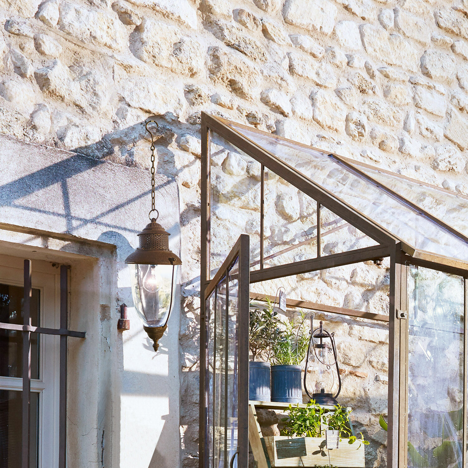 Glashaus mit Metallrahmen und Pflanzen vor einer Steinmauer im Sonnenlicht