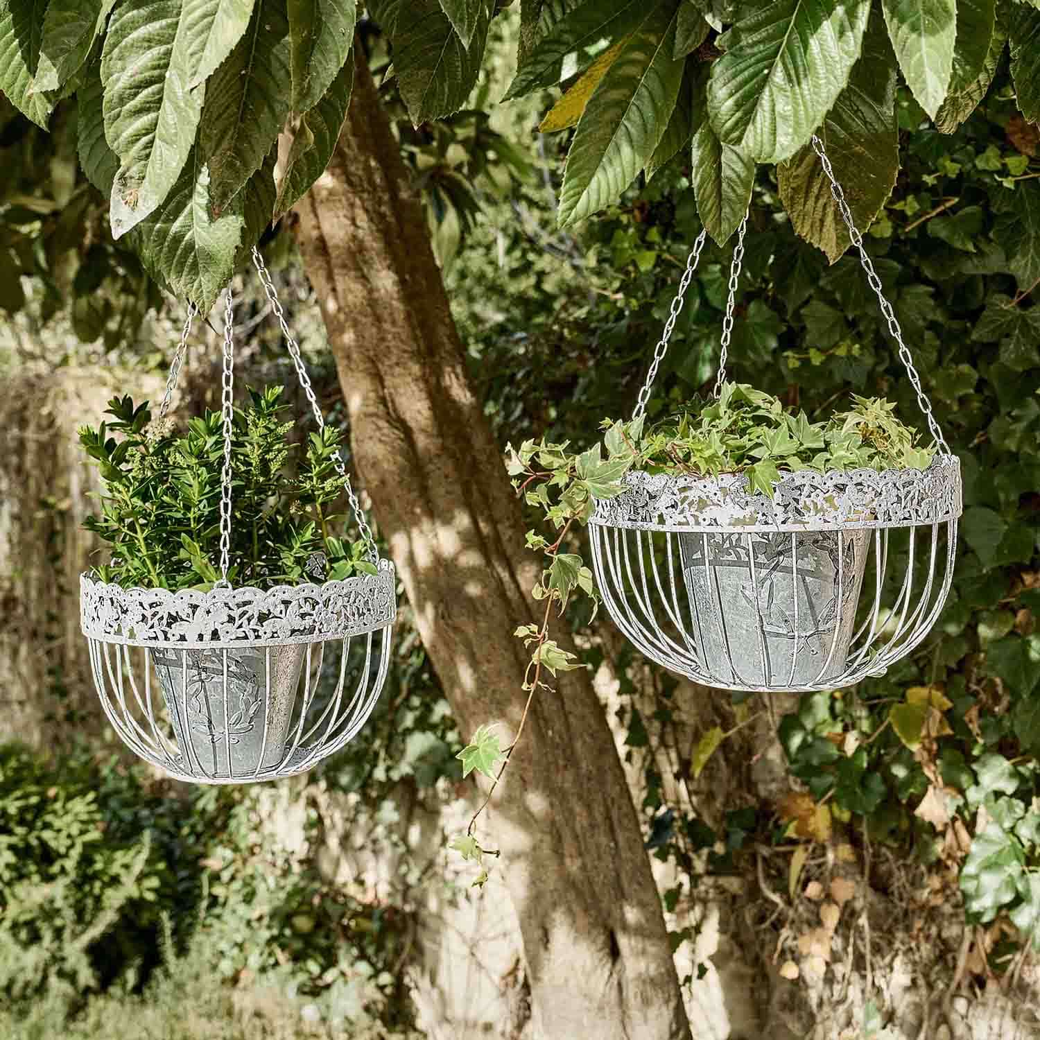 Zwei hängende Metallpflanzkörbe in Silber mit dekorativem Muster und grünen Pflanzen im Garten