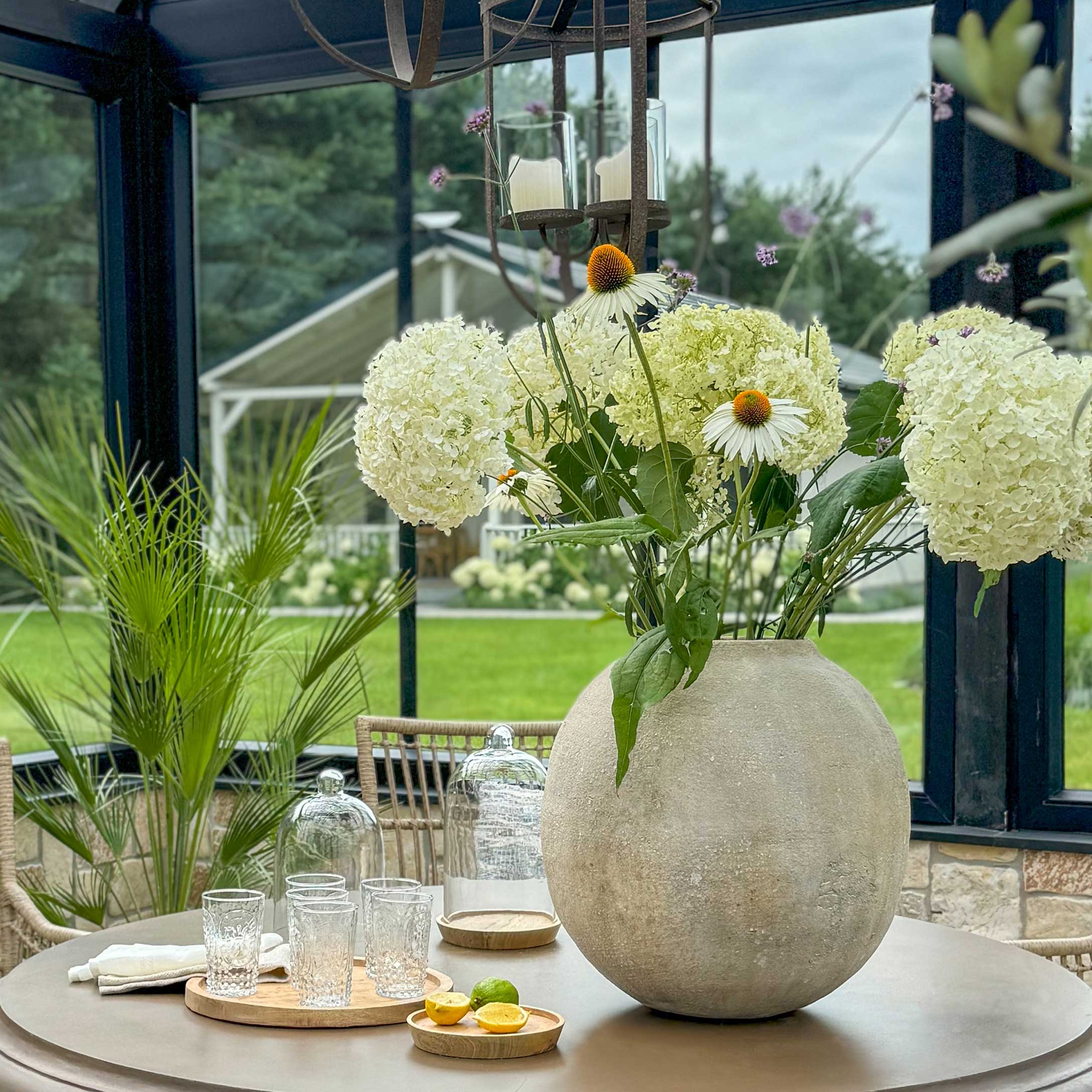 Große runde Betonvase mit weißen Blumen auf Tisch im Wintergarten mit Dekoration und Glas
