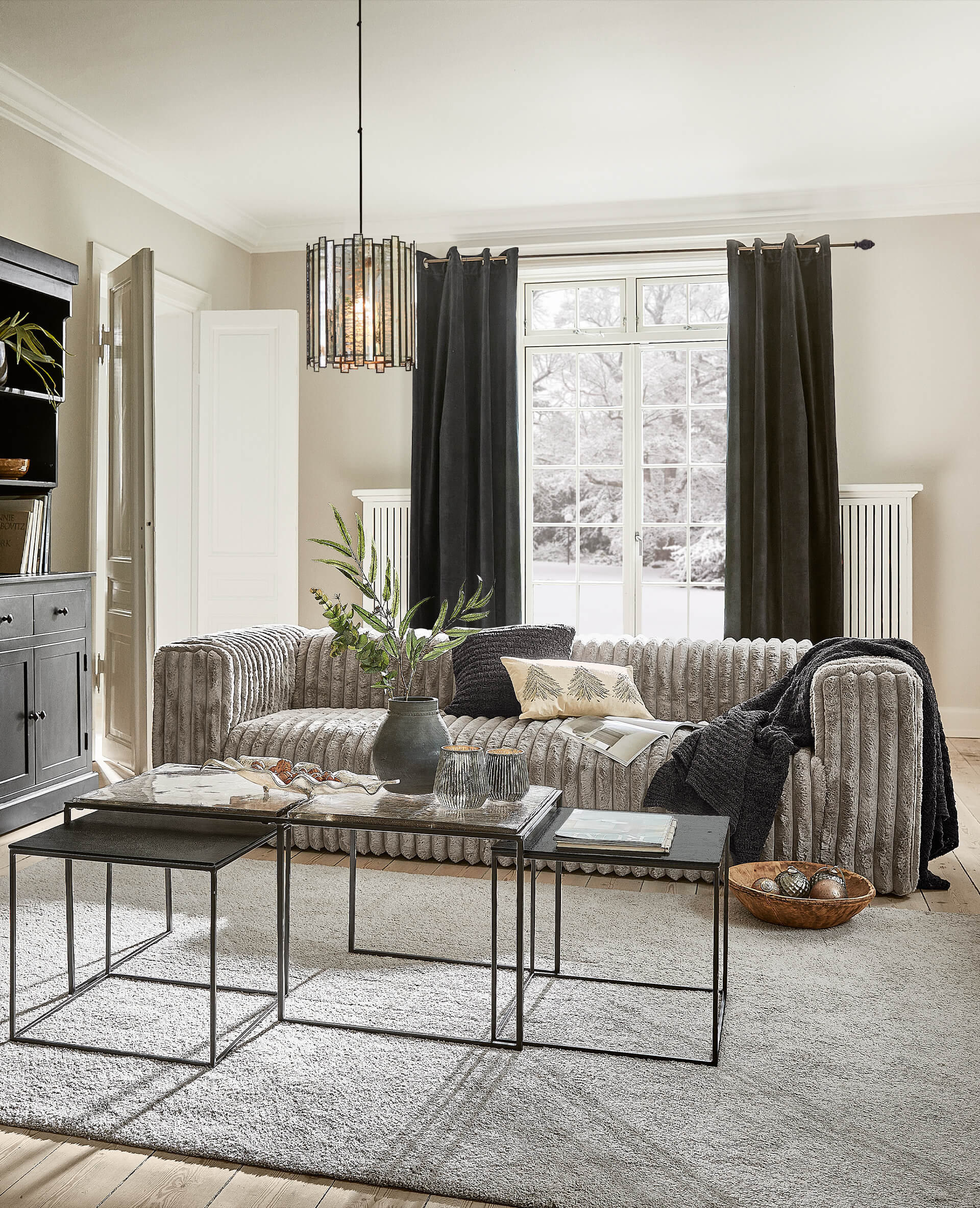 Moderne graue Couch aus weichem Material im hellen Wohnzimmer mit schwarzen Vorhängen und Metall-Couchtischen