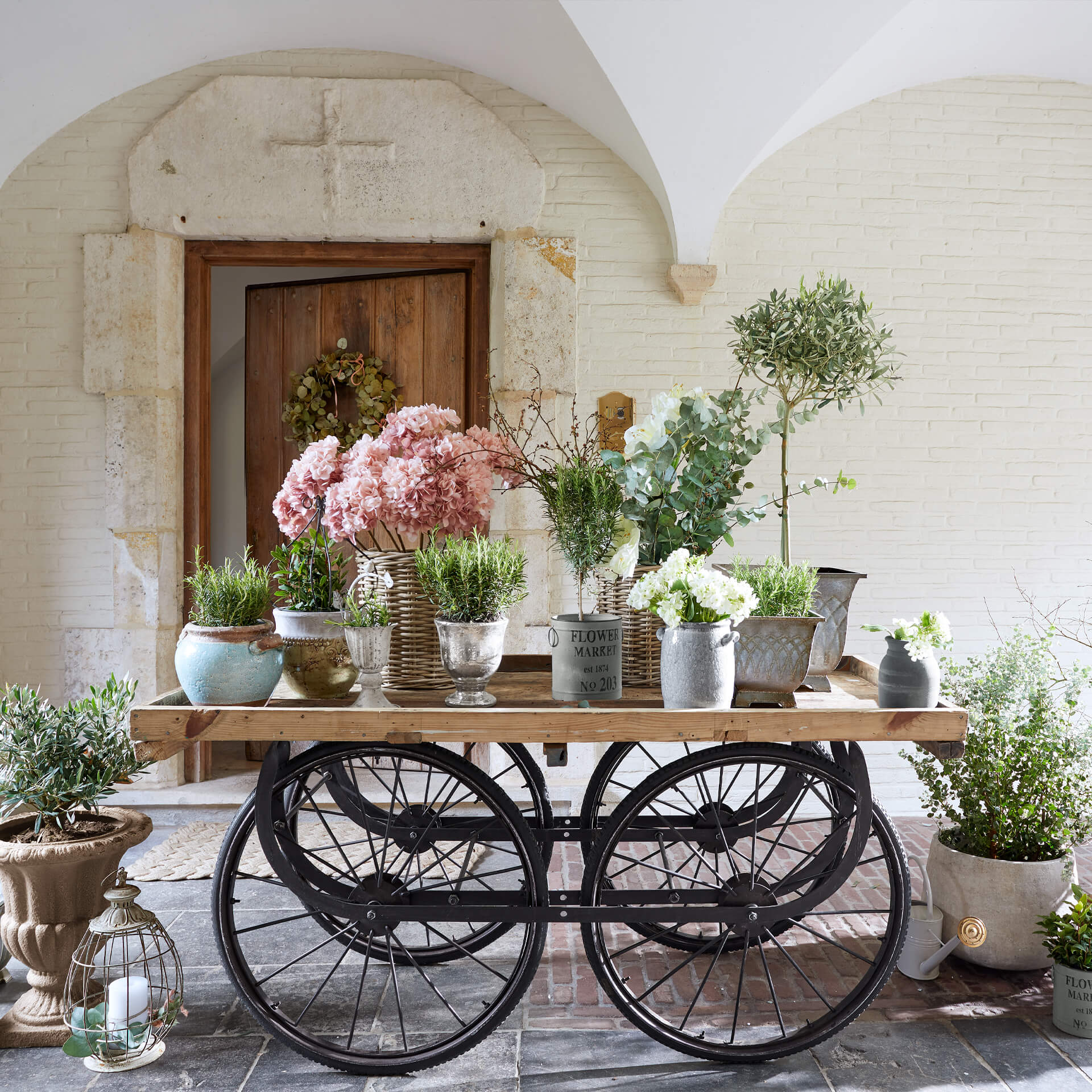Holzwagen mit schwarzen Metallrädern, verschiedenen Metalltöpfen und rosa sowie weißen Dekoblumen