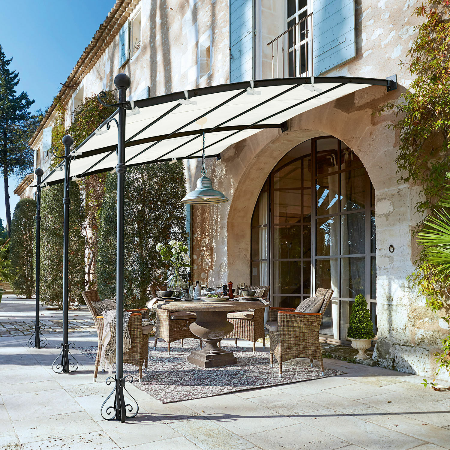 Pergola Kattles Gartenmöbel-Set aus Rattan unter weißem Vordach auf steinerner Terrasse vor einem Steinhaus