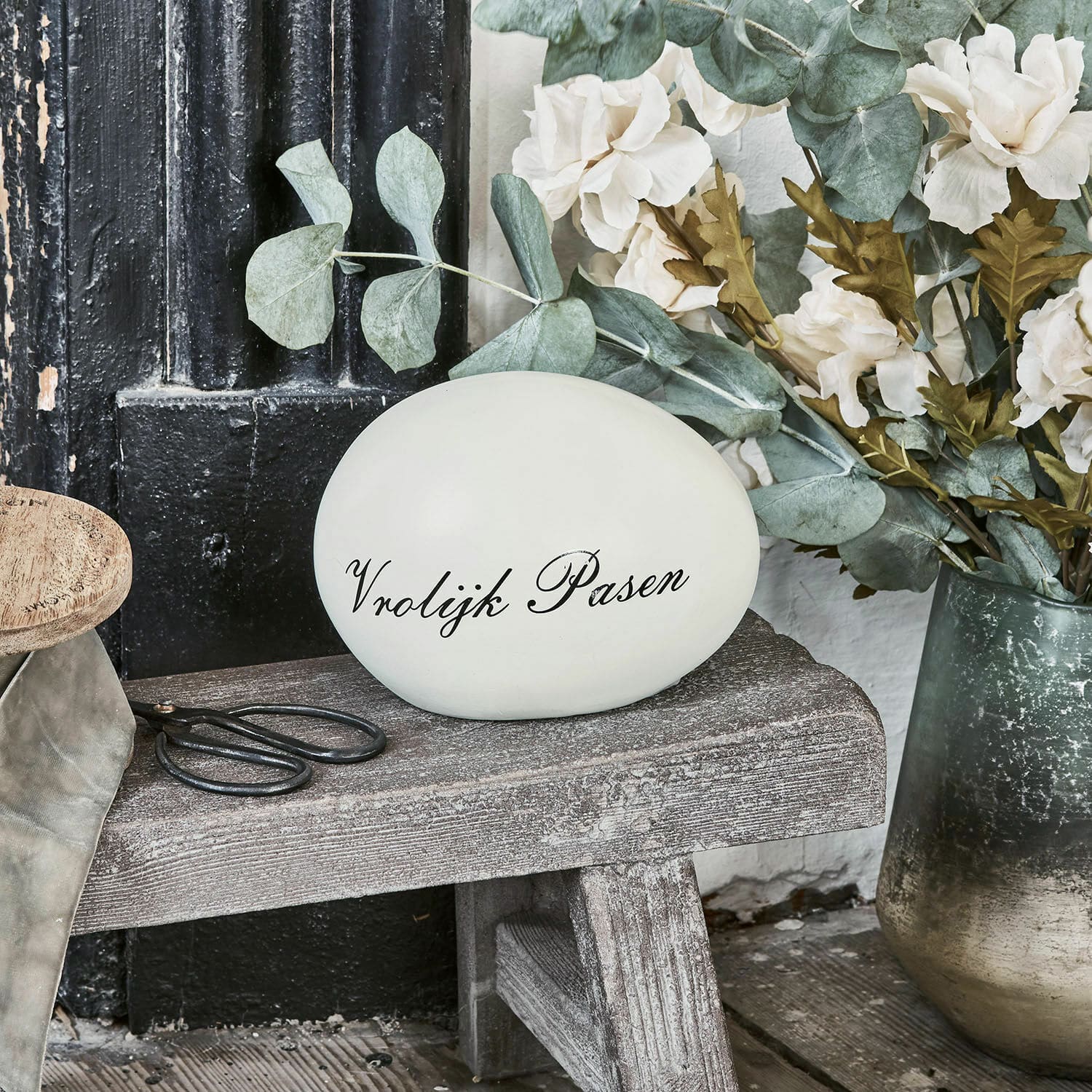 Deko-Ei VROLIJK PASEN Dekoratives weißes Ei mit Schriftzug Vrolijk Pasen auf Holzbank neben Blumen und Schere
