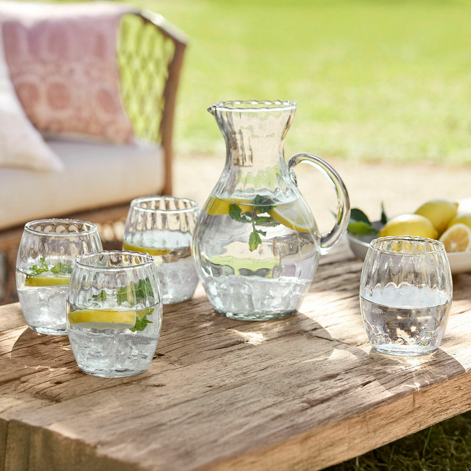 Glaskrug und vier Gläser mit Wasser, Zitrone und Minze auf rustikalem Holztisch im Garten