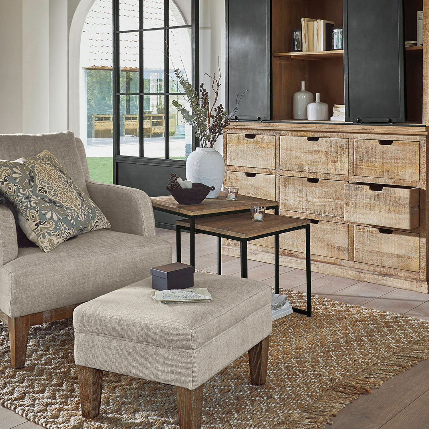 Gemütliches Wohnzimmer mit beigem Stoffsessel und Hocker, hellem Holzkommode und Metall-Couchtischen