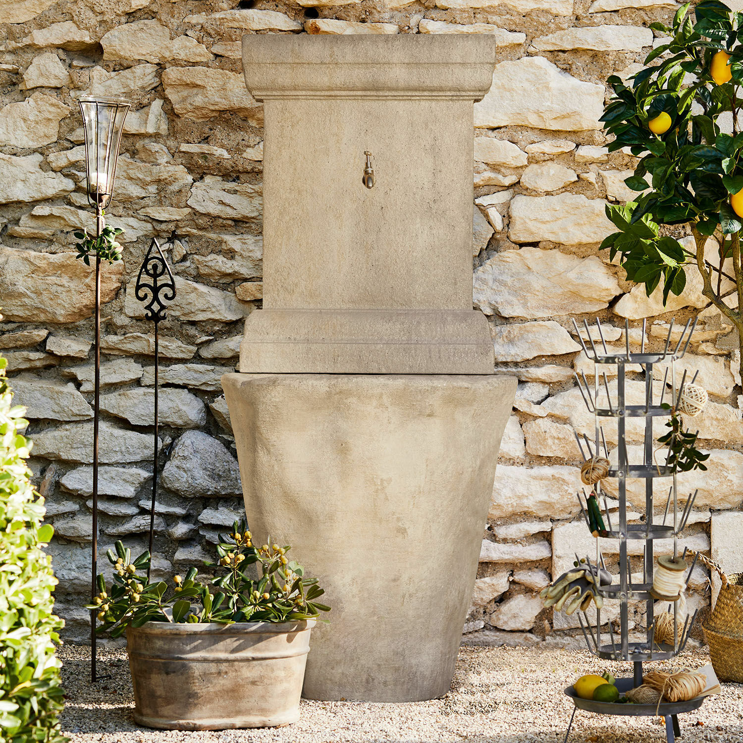 Großer Gartenbrunnen aus beigem Stein vor einer Steinmauer mit Pflanzen im Hintergrund