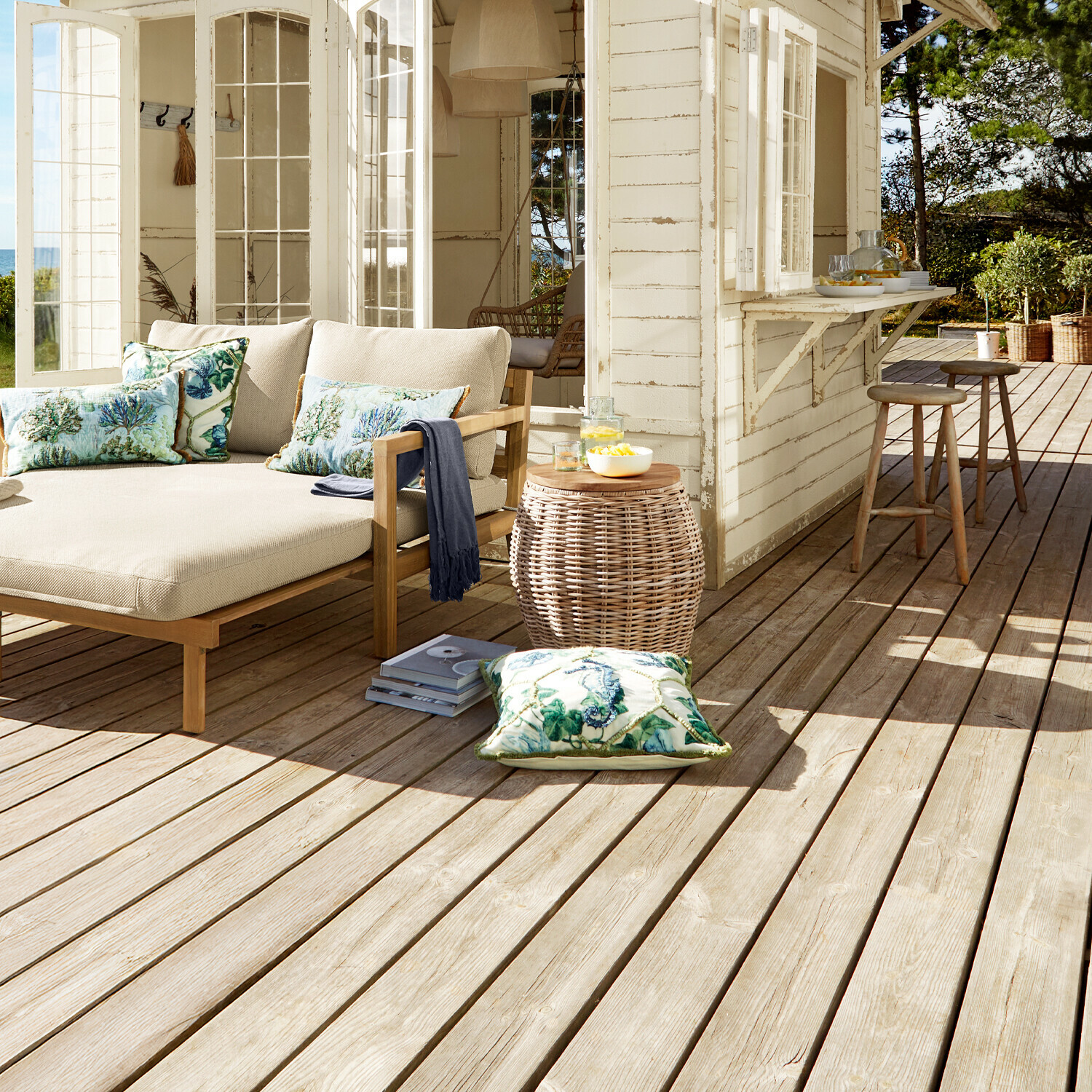 Gemütliche Terrasse mit beige Sofa auf Holzrahmen und Kissen mit maritimem Pflanzenmuster