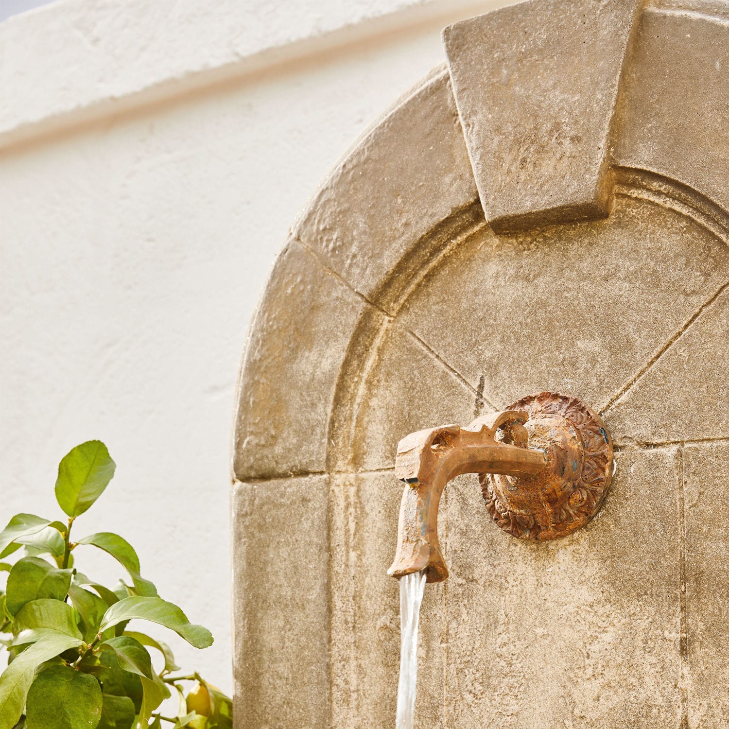 Rostiger Messing-Wasserhahn an Steinmauer mit fließendem Wasser und grünen Blättern daneben