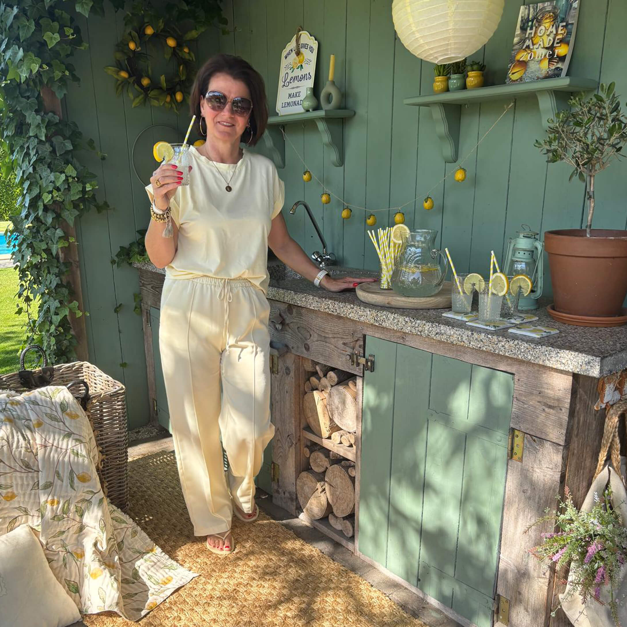 Frau in cremefarbenem Outfit steht an grüner Außenküche mit Limonade und Zitronendekor