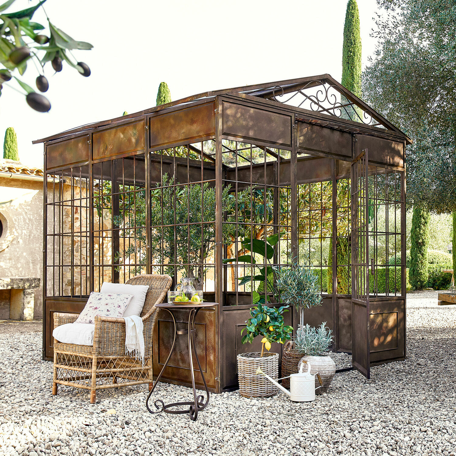 Pavillon Essone Rustikale Metall-Orangerie auf Kiesboden mit Rattansessel und dekorativen Pflanzen im Garten