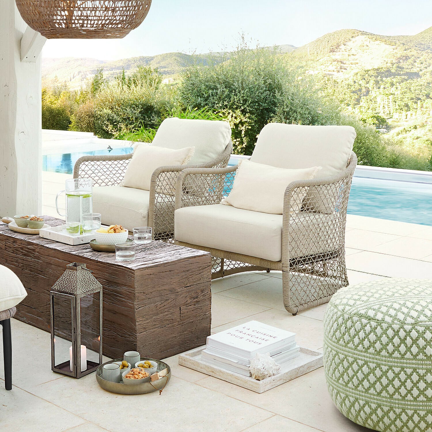 Zwei beige Sessel aus geflochtenem Material auf Terrasse am Pool mit Holztisch und grünem Pouf