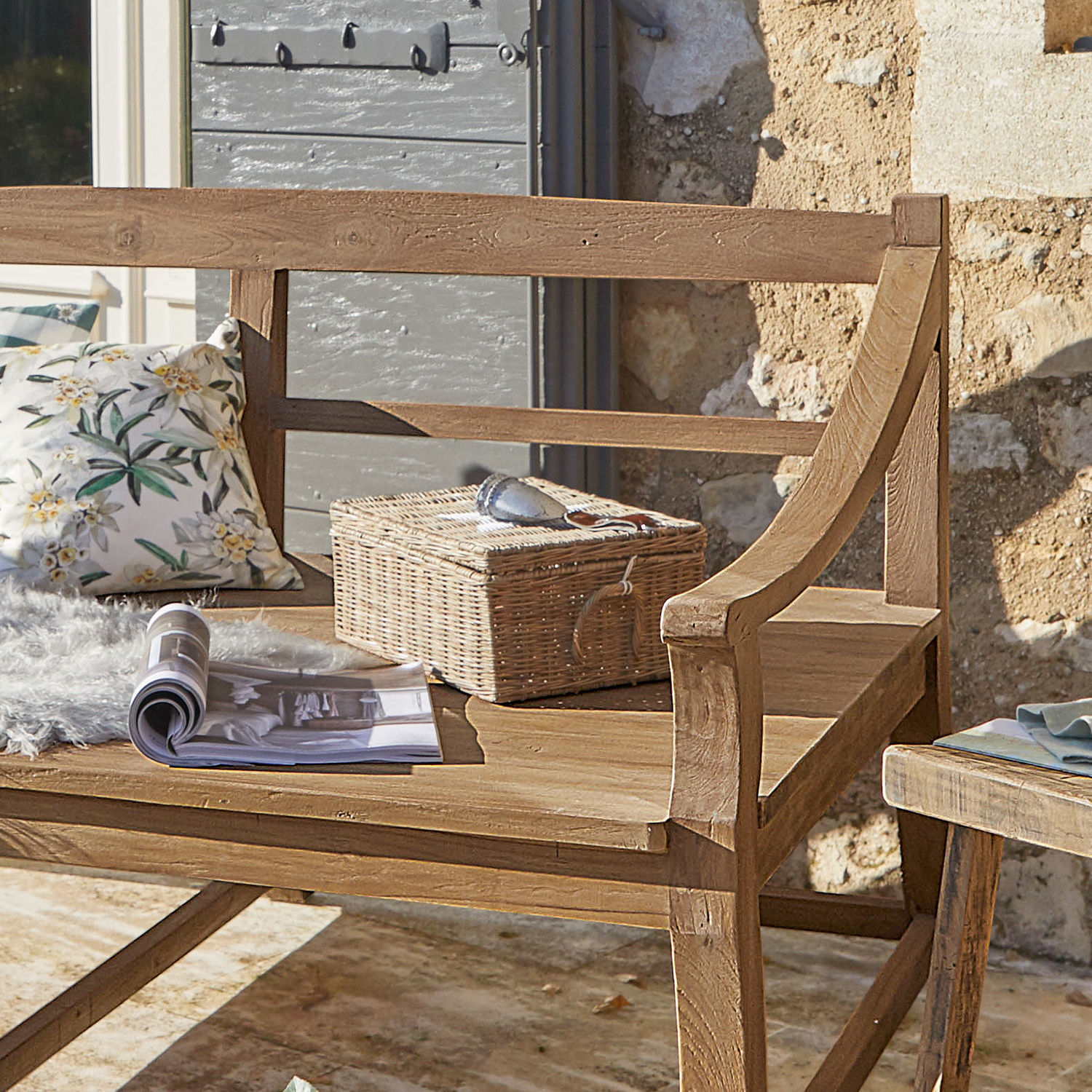 Holzbank mit Korb aus Weide und floralem Kissen auf sonniger Terrasse