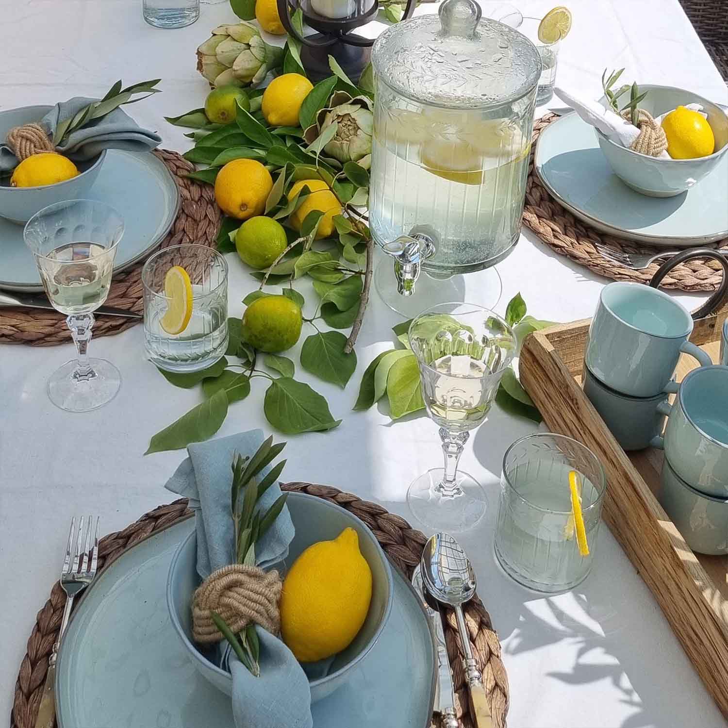 Sommerlicher Tisch mit blauer Keramik, Zitronen und grünen Blättern auf weißem Tischtuch, Tischdeko