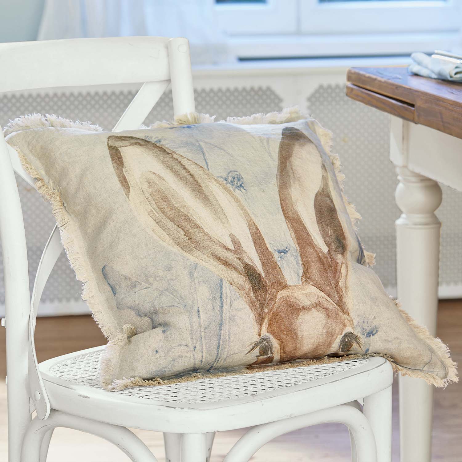 Beiges Leinenkissen mit Hasenohren-Motiv und Fransen auf weißem Stuhl im Wohnzimmer