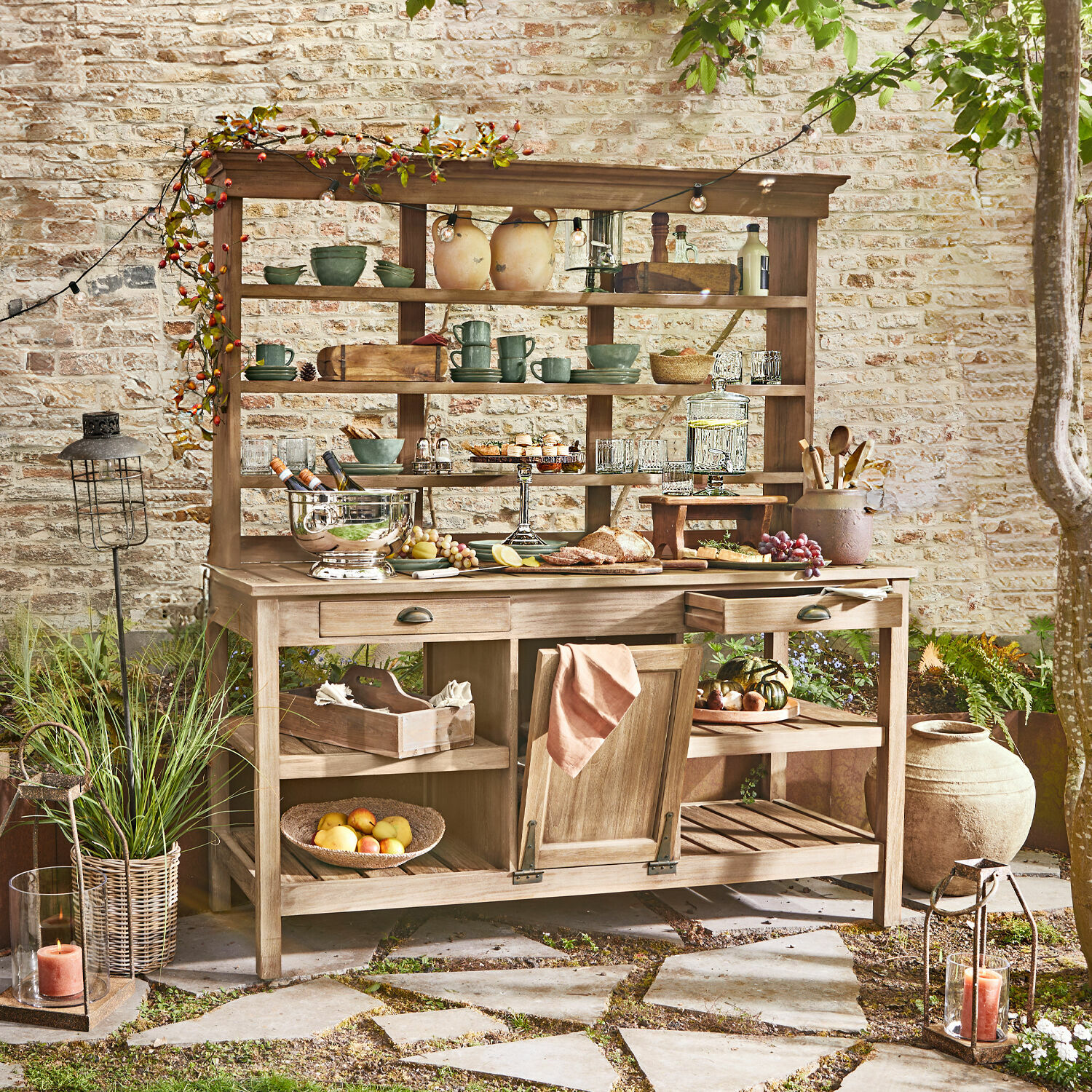 Gartenschrank Fallon Holz-Gartenmöbel mit Regalen und Schubladen, dekoriert mit Geschirr und Speisen auf Terrasse