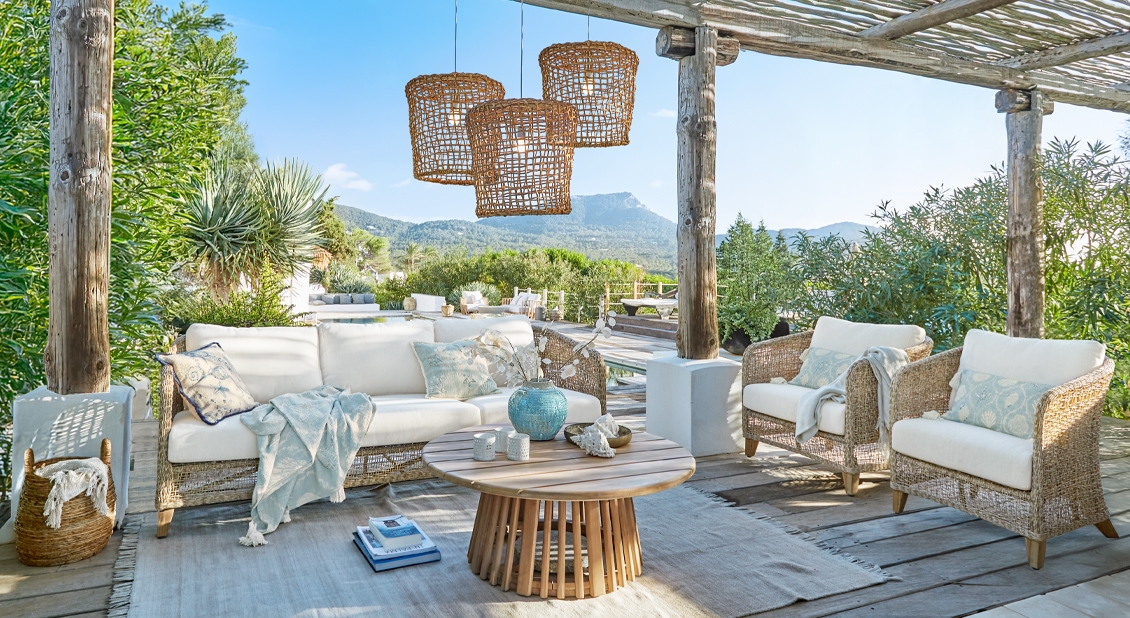 Outdoor-Rattansitzgruppe in Natur mit weißen Polstern und rundem Holztisch auf Terrasse mit Bergblick