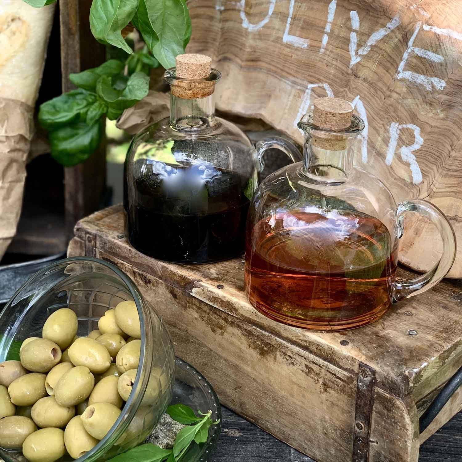 Glasflaschen mit Korken gefüllt mit Öl und Essig auf Holzkiste neben grünen Oliven und Basilikumblättern