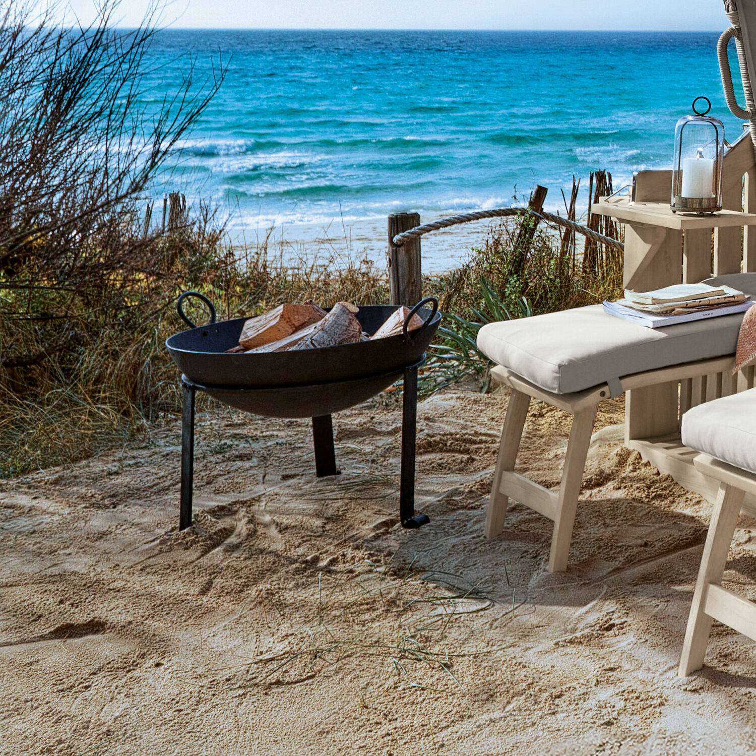 Schwarzes Metall-Feuerkorb mit Holz am Strand neben hellen Gartenmöbeln und Meer