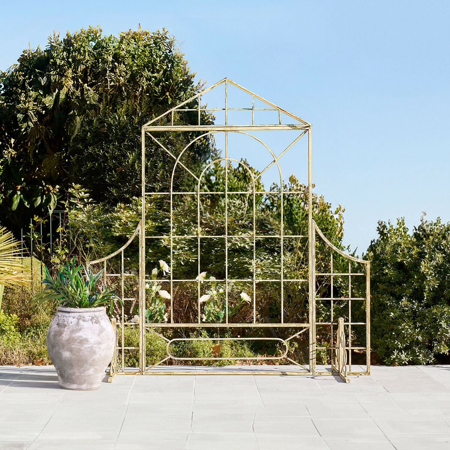 Goldfarbenes Metall-Gartenrankgitter in Hausform auf Terrasse mit Pflanzgefäß und Grün im Hintergrund