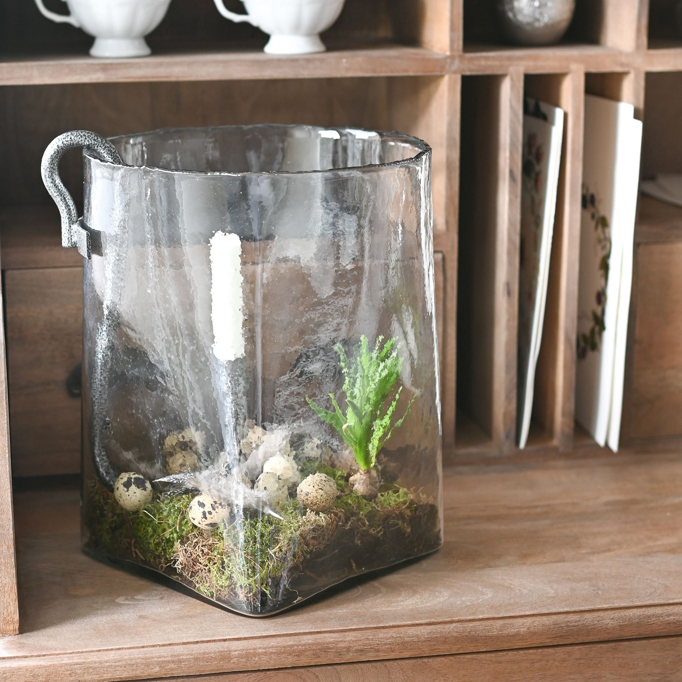 Transparenter Glasbehälter mit Griff, dekoriert mit Moos und Wachteleiern auf einem Holztisch