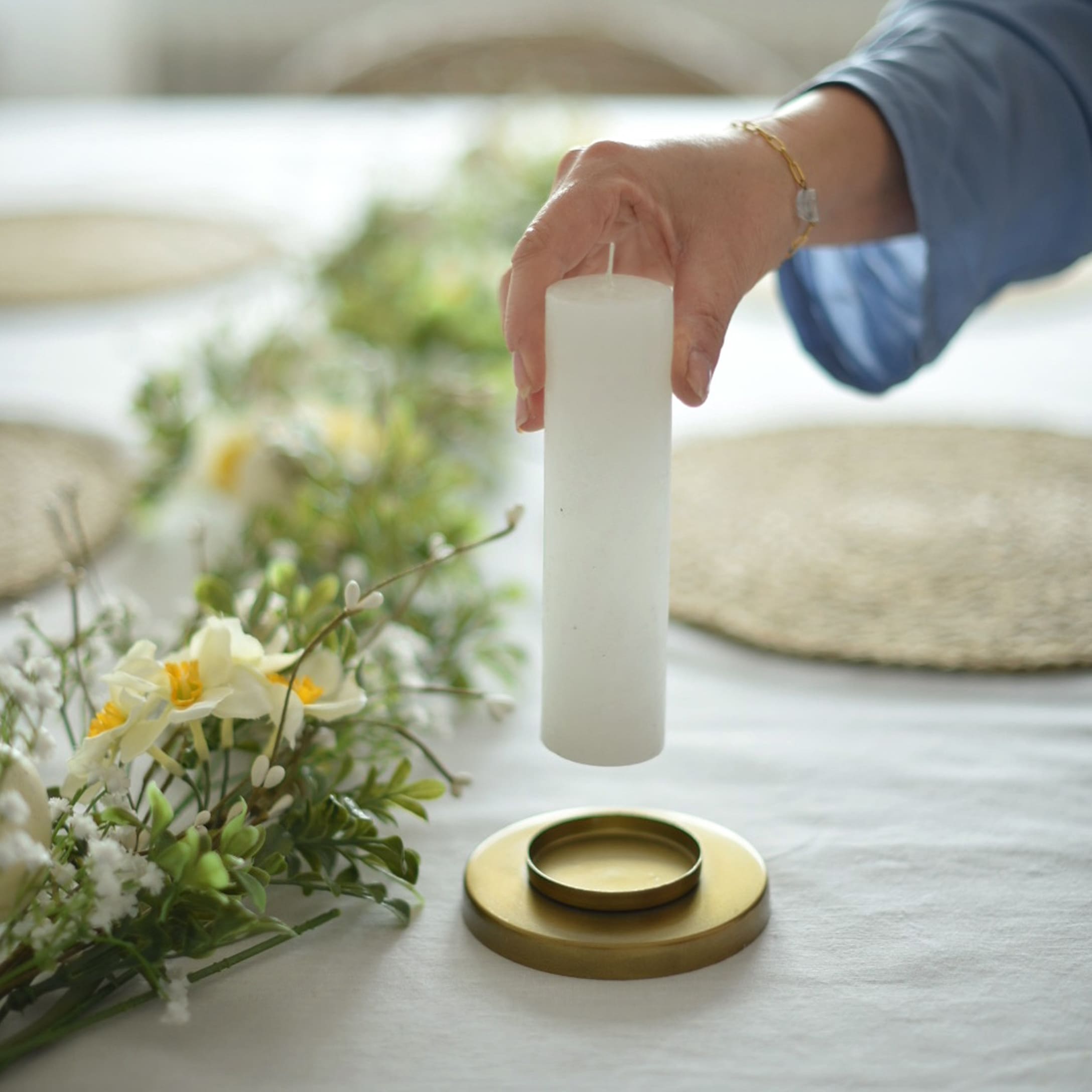 Weiße Stumpenkerze über goldenem Kerzenhalter mit Blumenarrangement auf weißem Tischtuch