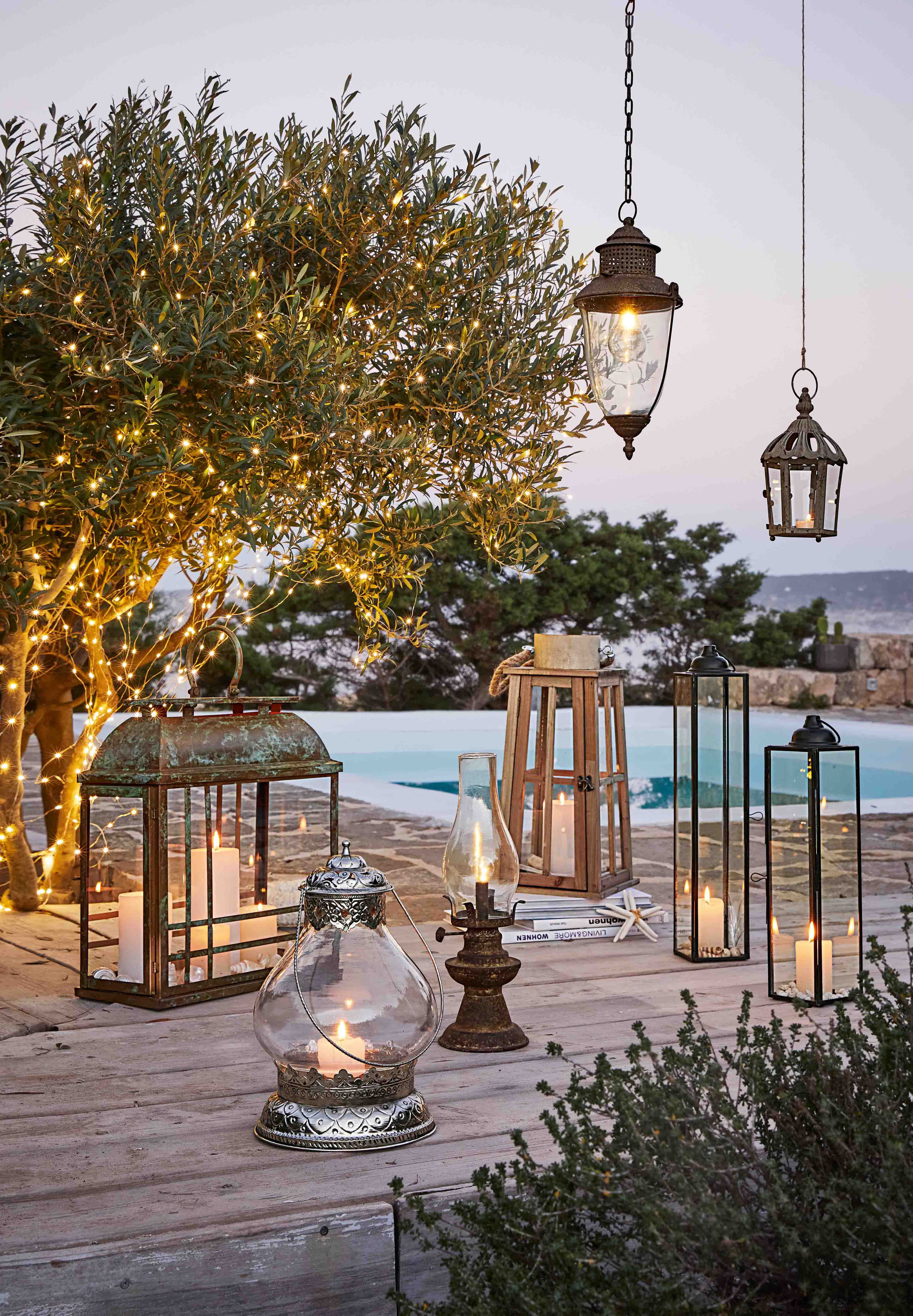 Verschiedene Laternen und Kerzen auf Holzterrasse mit Baum, der mit LED-Lichterketten geschmückt ist