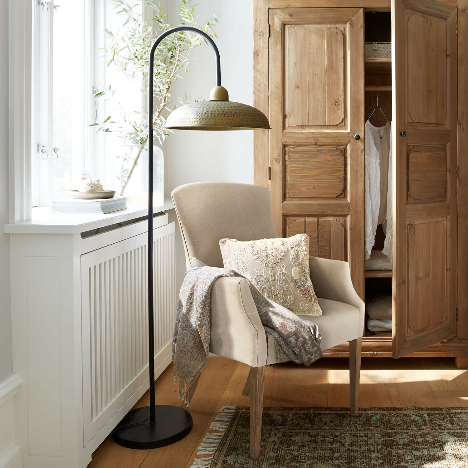 Stehlampe Moorford Beiger Sessel mit Holzbeinen, dekorativem Kissen und Decke, daneben schwarze Stehlampe mit messingfarbenem Schirm