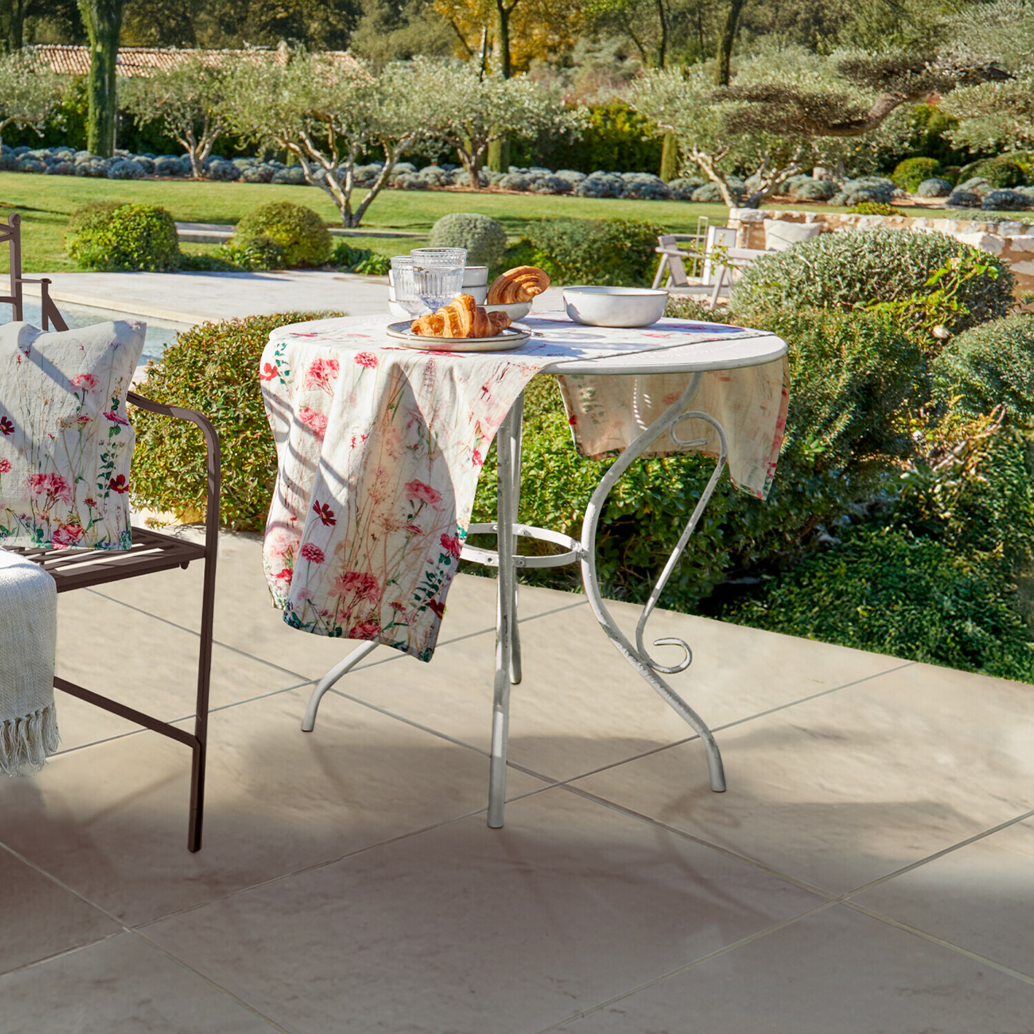 Tisch Aube Runder weißer Metall-Gartentisch mit floralem Tischtuch und Gebäck auf Terrasse mit Gartenblick