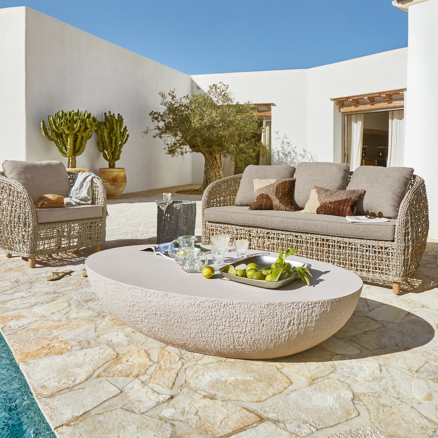Gartenmöbel-Set mit beigem Steintisch und grauem Polstersofa aus Rattan auf Natursteinboden