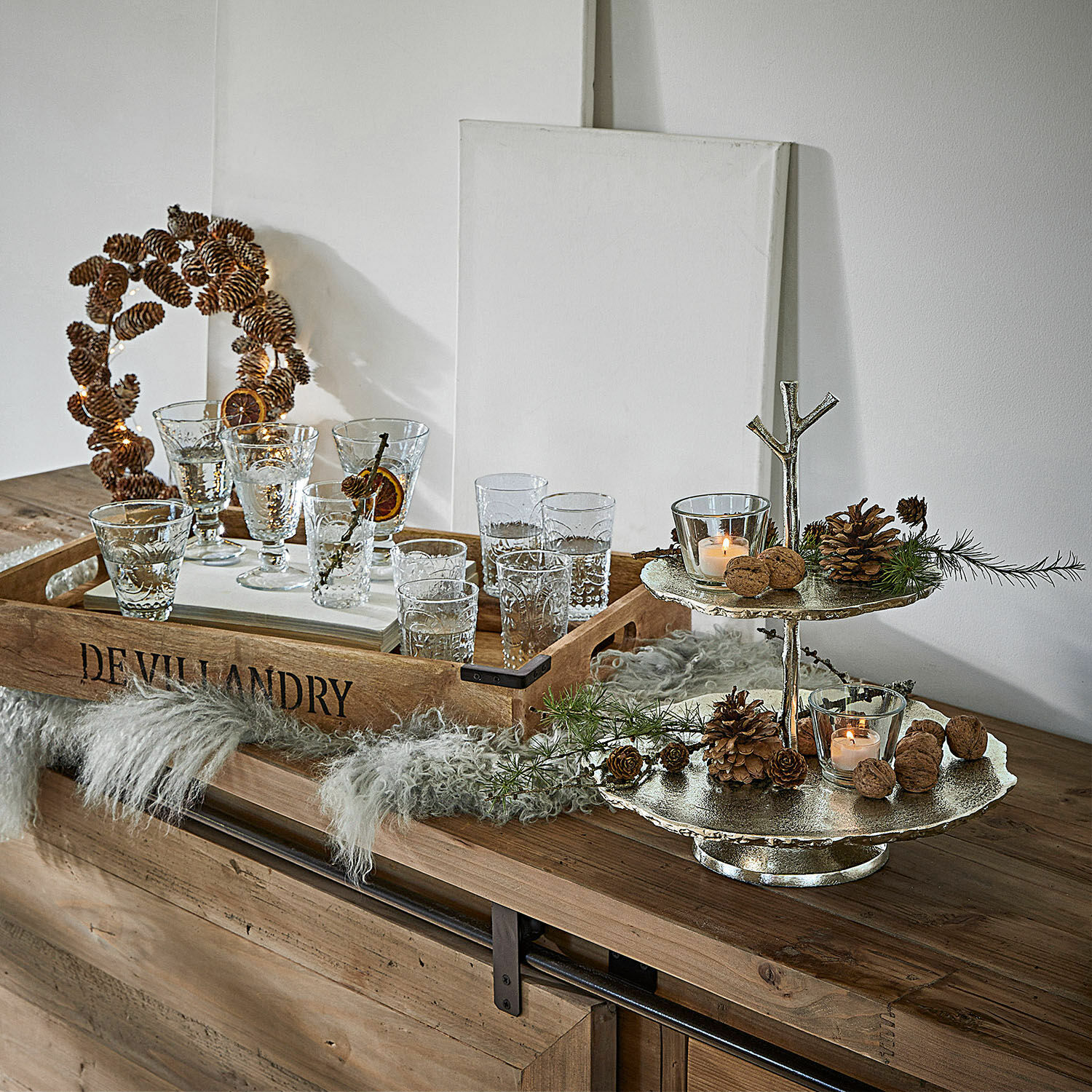 Zweistöckige Metall-Etagere mit Tannenzapfen und Kerzen auf Holzkommode mit Tablett und Gläsern