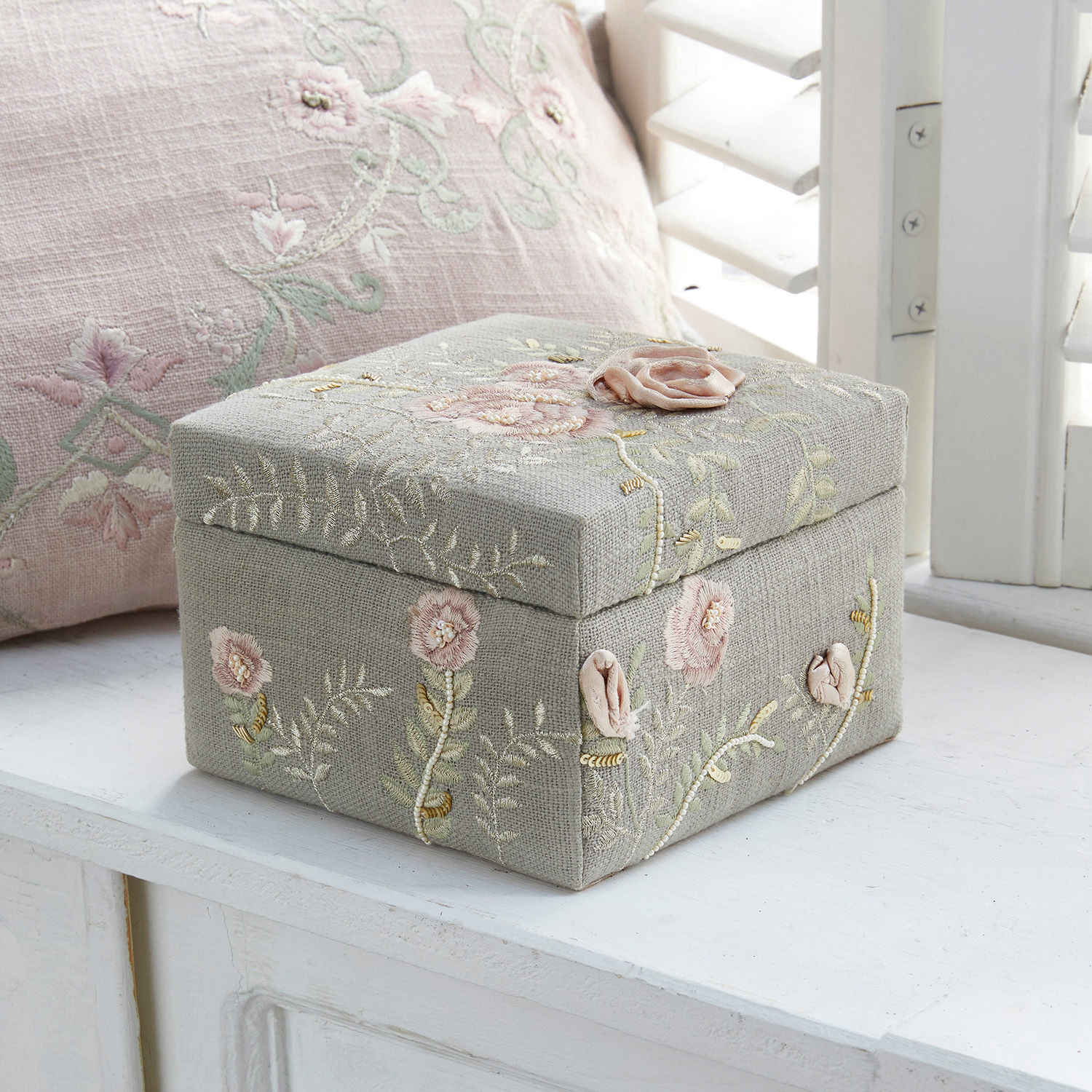 Graue Leinenbox mit pastellfarbenem Blumenstickerei auf weißem Fensterbrett