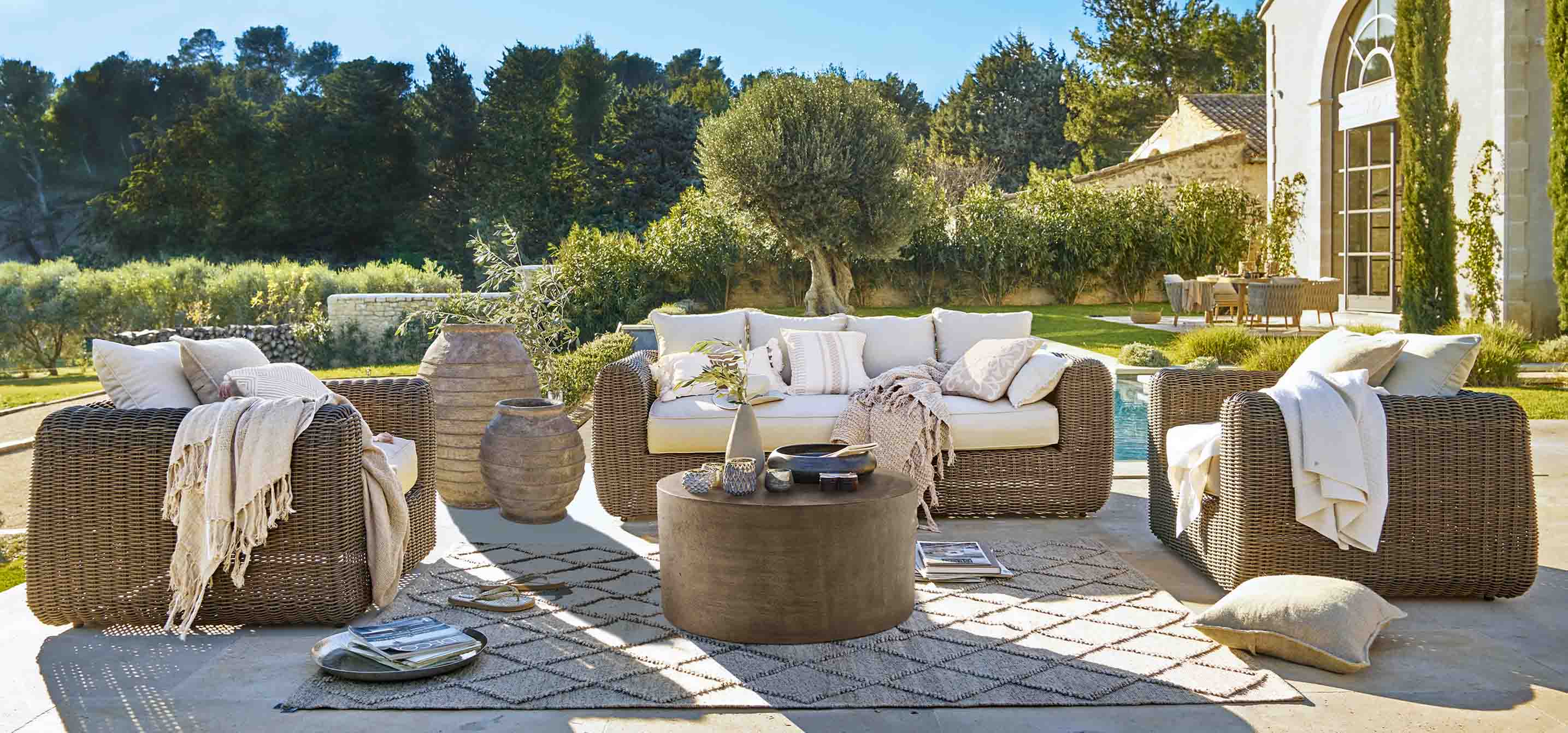 Braunes Rattan-Gartenmöbel-Set mit cremefarbenen Kissen und rundem Couchtisch auf sonniger Terrasse