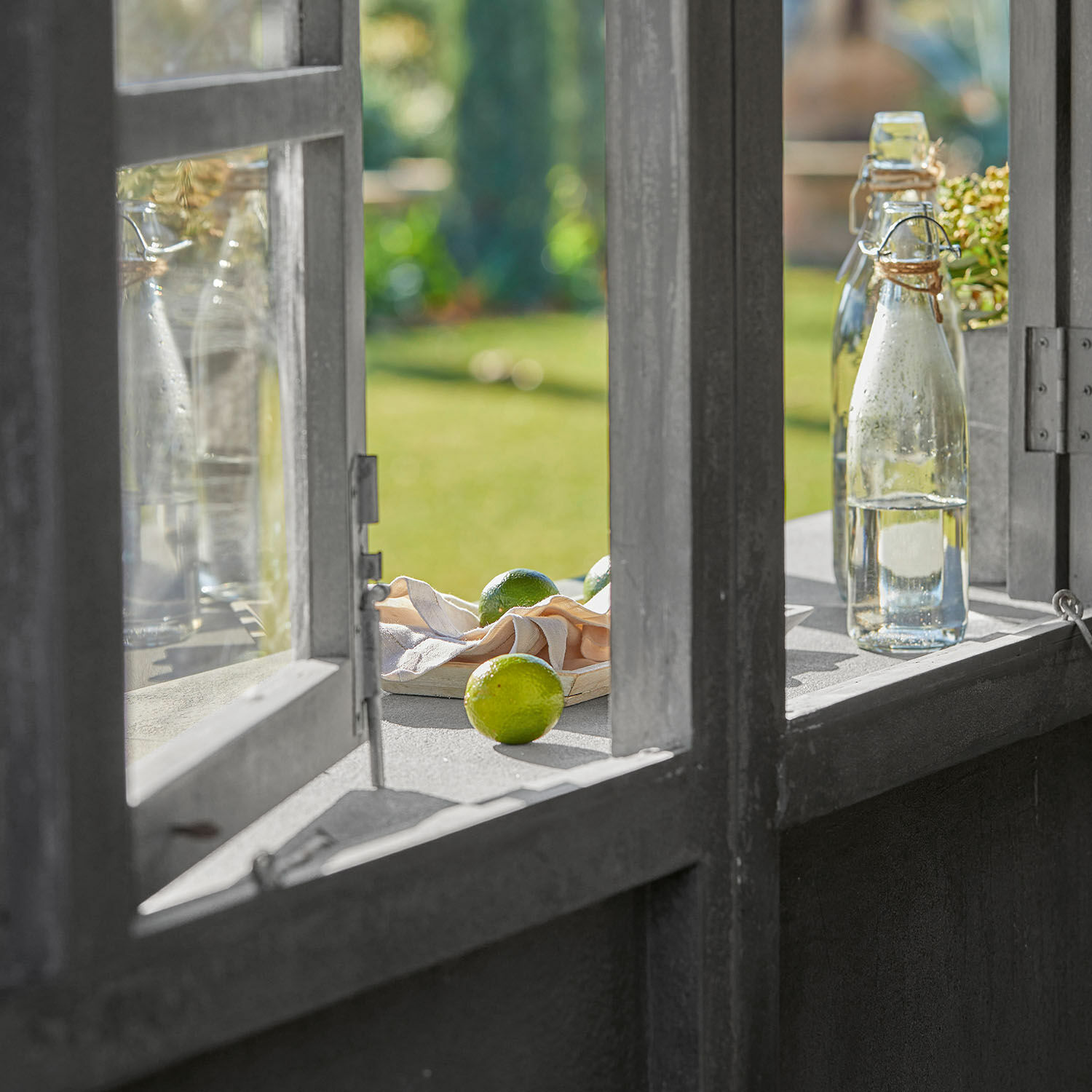 Graues offenes Fenster mit Wasserflaschen und Limetten auf dem Sims, grüner Garten im Hintergrund