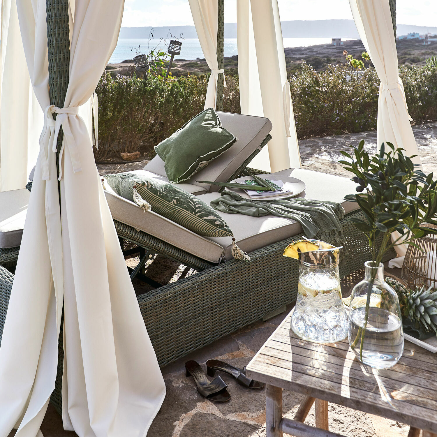 Grüne Rattan-Liege mit cremefarbenen Polstern und grünen Kissen auf Terrasse mit Meerblick