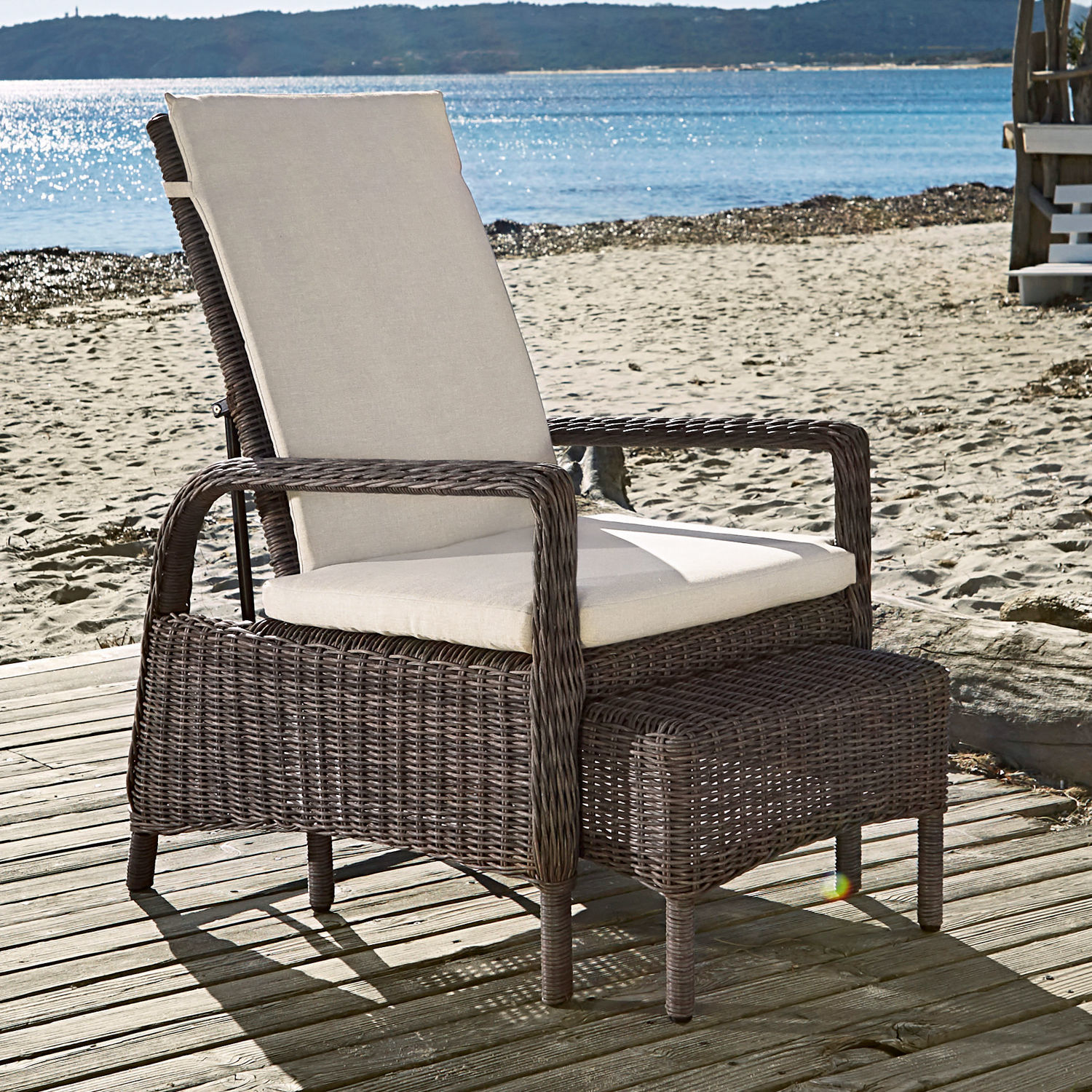 Gartenstuhl aus braunem Rattan mit cremefarbenem Polster und Fußhocker auf Holzterrasse am Strand