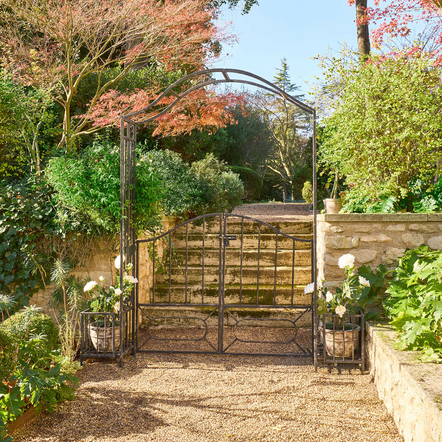 Schwarzes Metall-Gartentor mit Bogen und weißen Blumentöpfen auf sonnigem Kiesweg