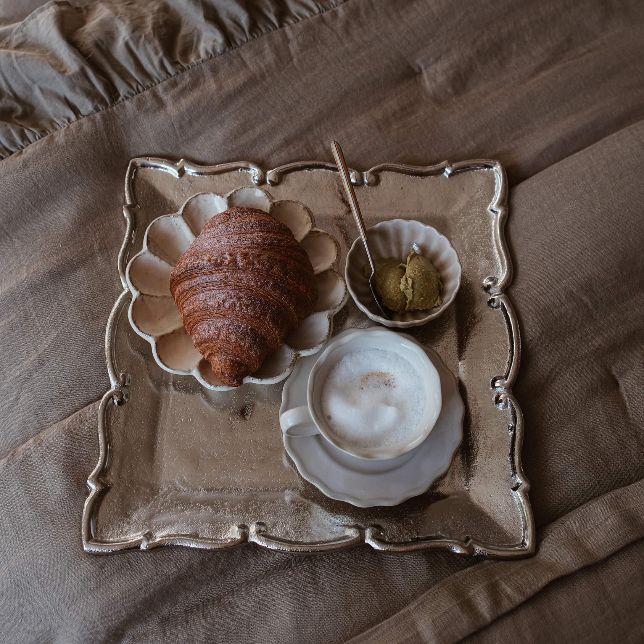 Tablett mit Croissant, Milchschaumkaffee und Aufstrich auf beigem Stoff im Slow-Living-Stil