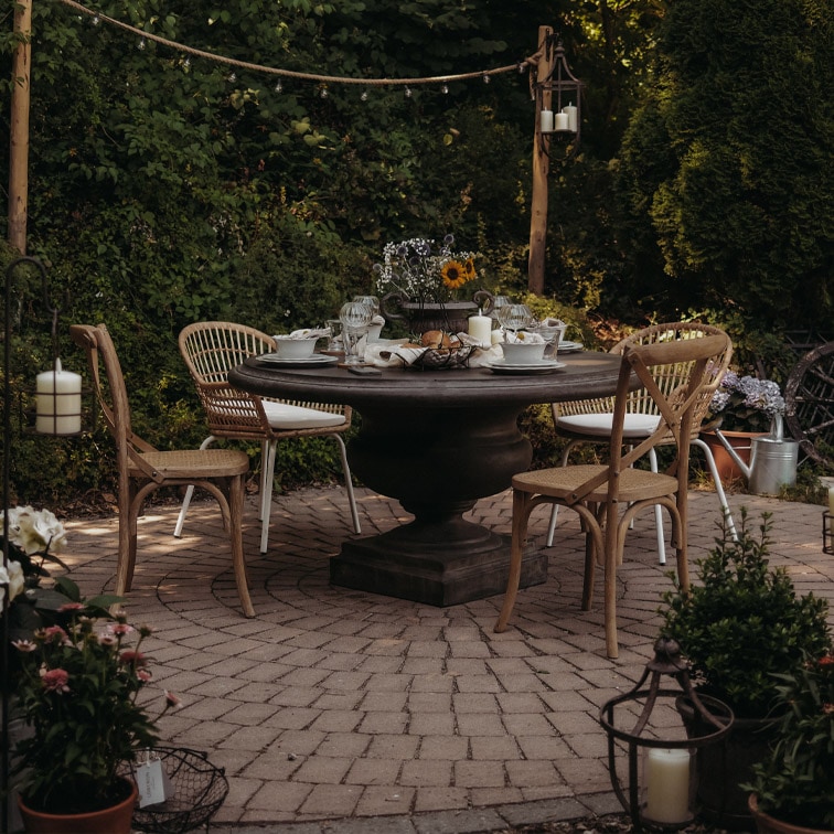 Runder Gartentisch aus dunklem Holz mit Rattanstühlen auf gepflasterter Terrasse mit Kerzen und Blumen dekoriert