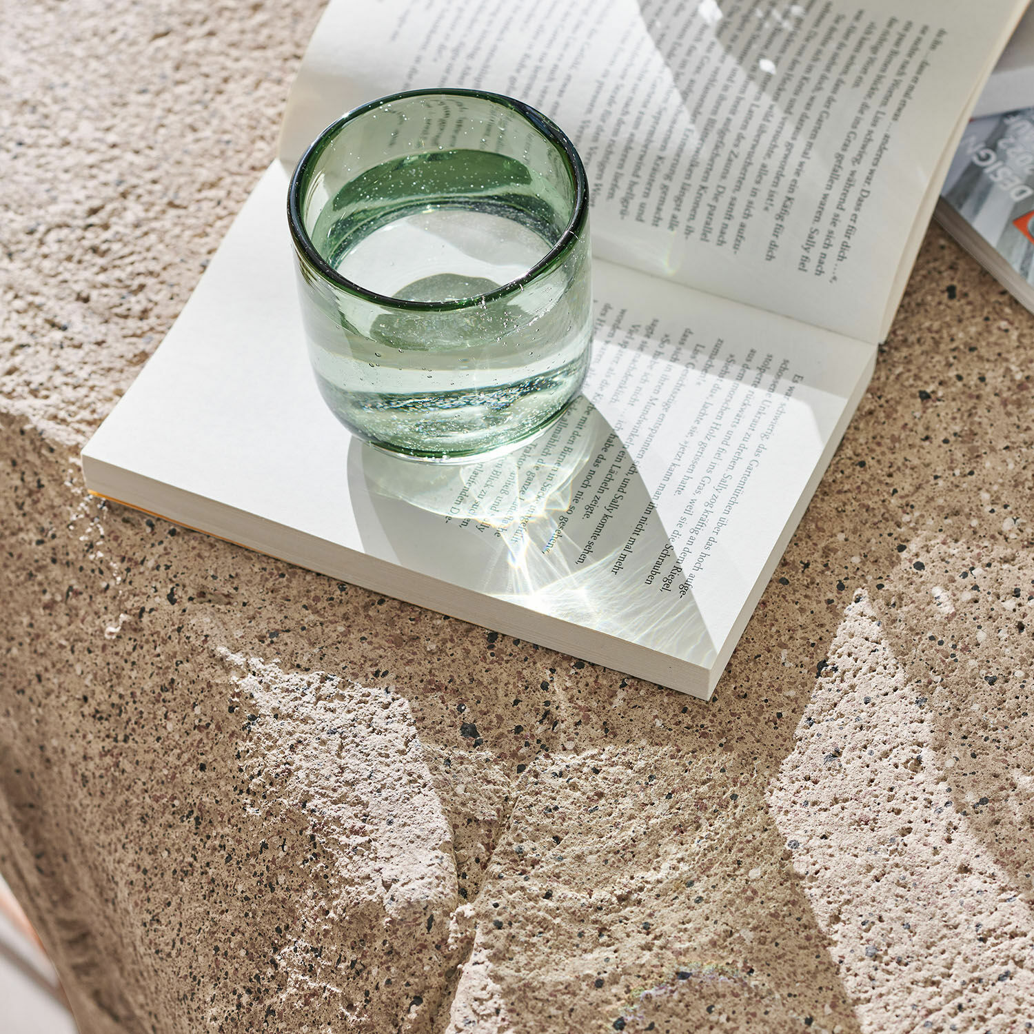 Grünes Glas mit Wasser auf offenem Buch auf steinerner Oberfläche im Tageslicht