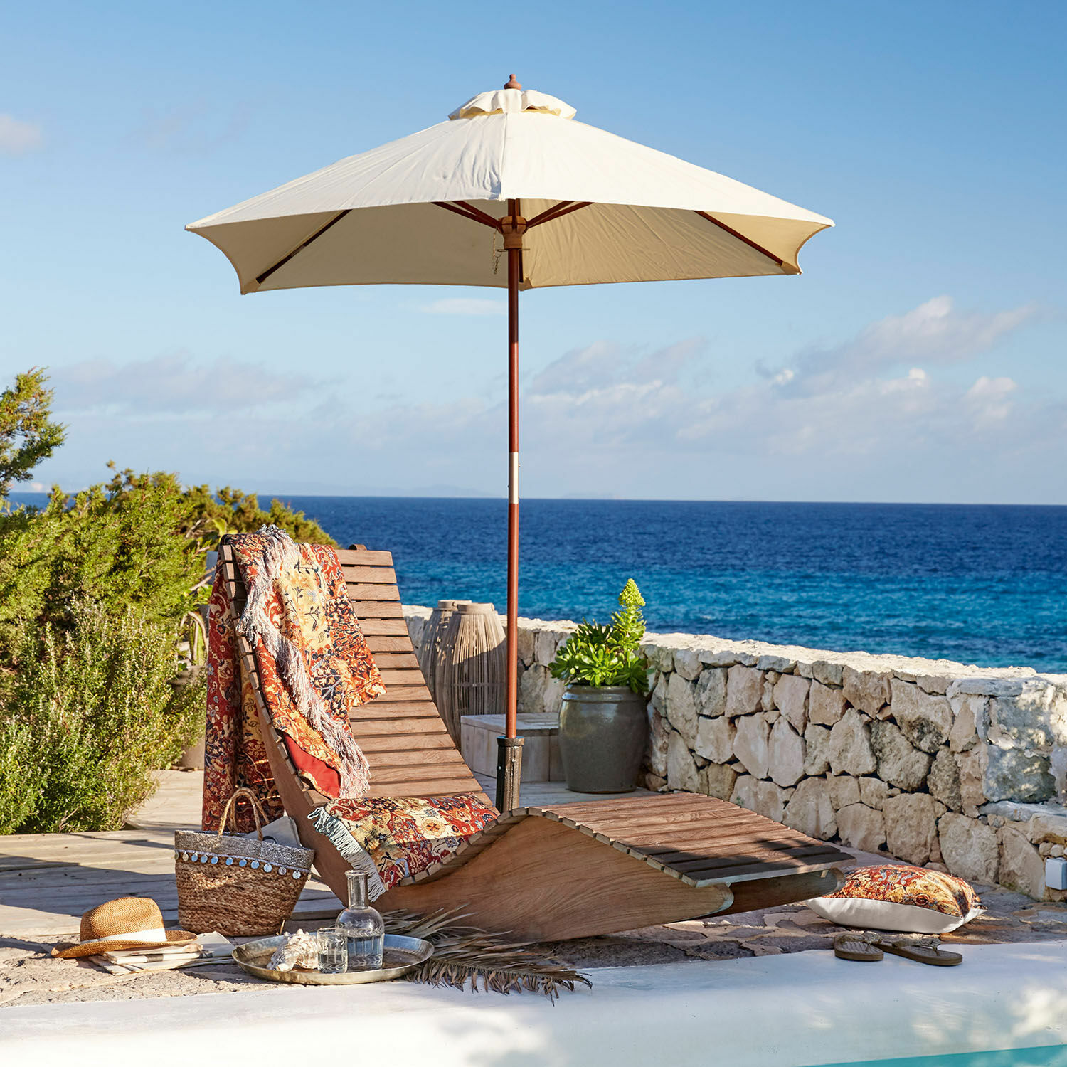 Lounger Wenham Holzliege mit cremefarbenem Sonnenschirm und buntem Plaid am Pool mit Meerblick