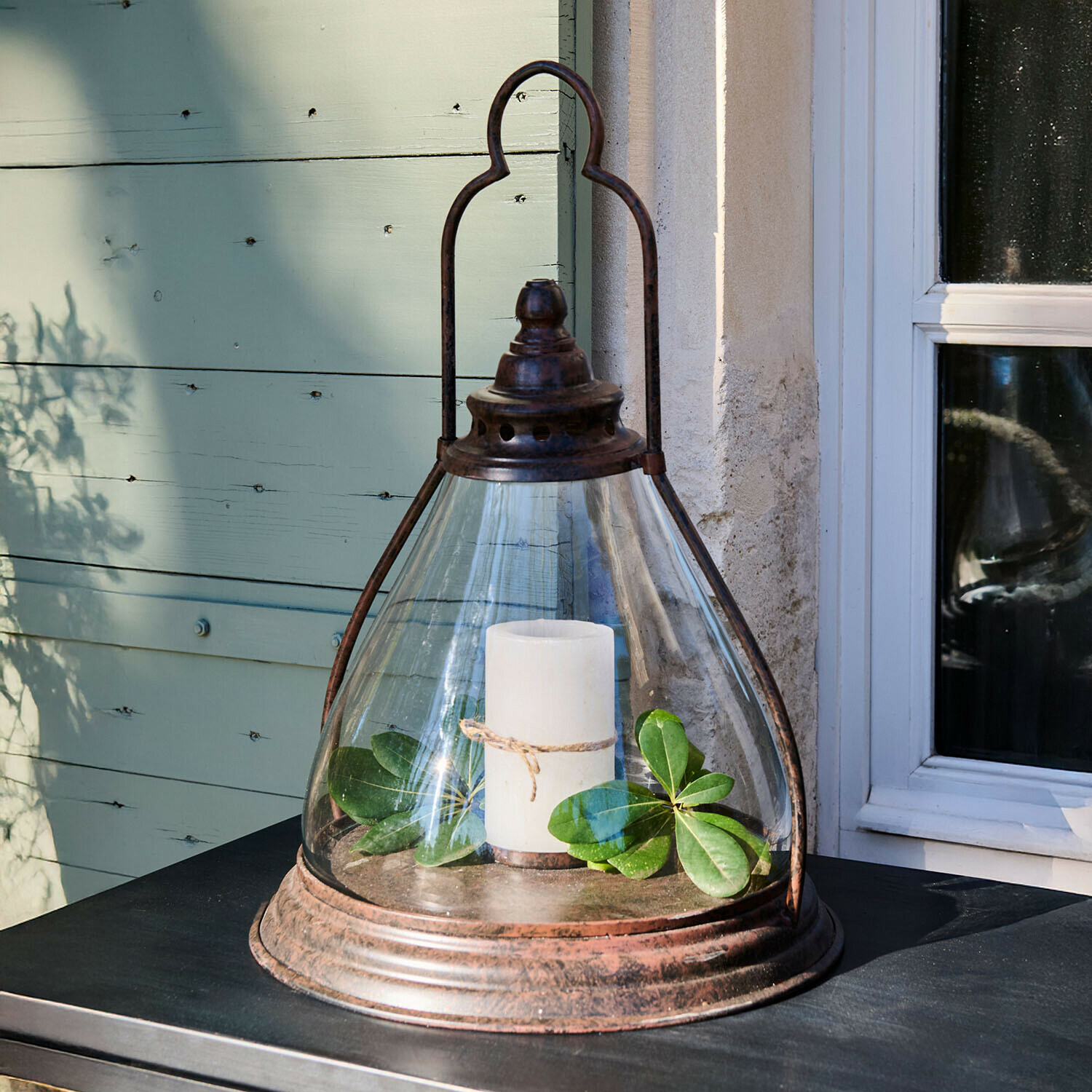 Glaslaterne mit rostigem Metallrahmen, weißer Kerze und grünen Blättern auf Tisch