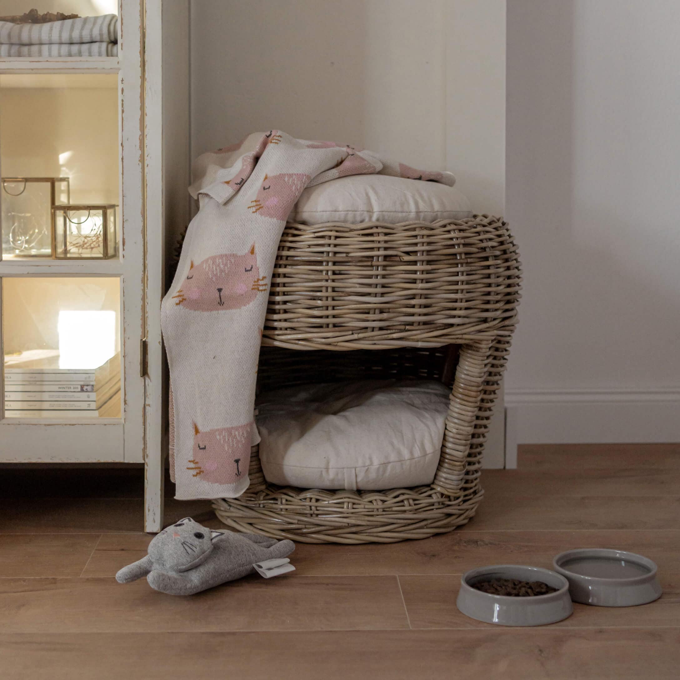 Zweistöckiges Katzenbett aus Rattan mit hellen Kissen, rosa Katzenmuster-Decke, Spielzeug und Futternäpfen