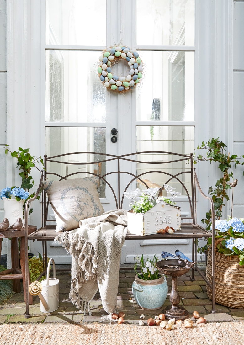 Metall-Gartenbank mit beiger Decke und Kissen, Blumenarrangements und pastellfarbener Eierkranz