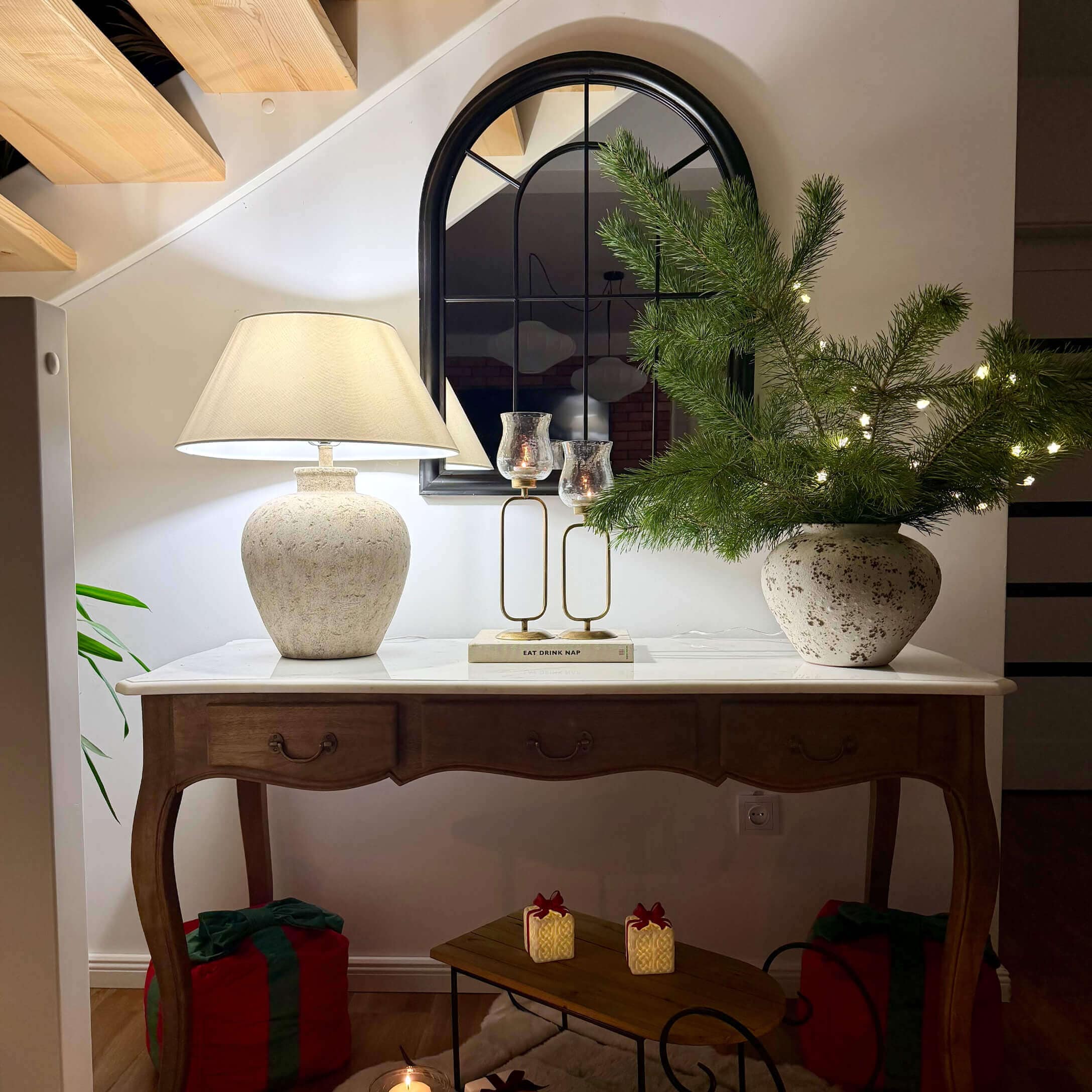 Holztisch mit weißer Platte, Keramiklampe, Spiegel und Weihnachtsdeko mit grünen Zweigen und Lichterkette