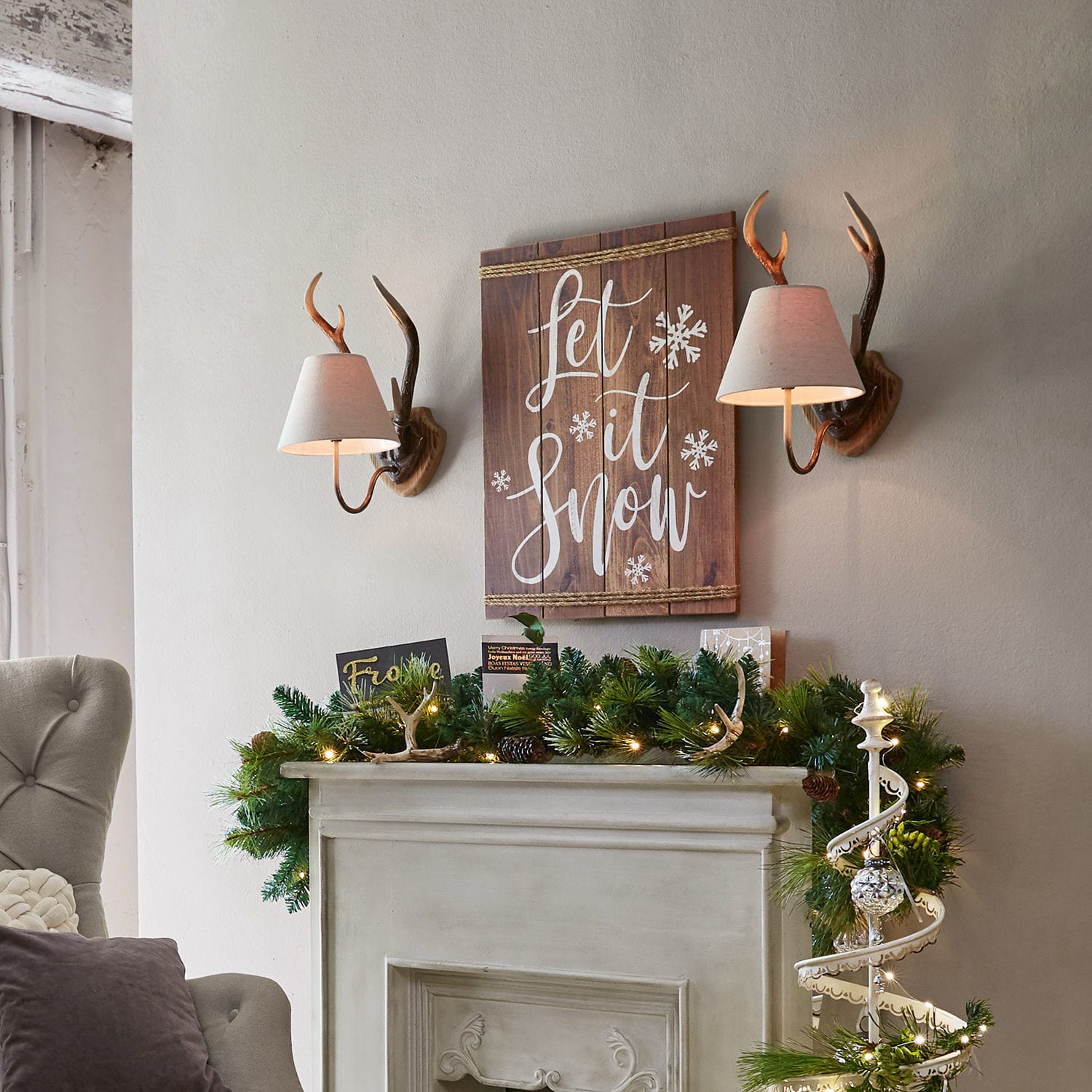 Wanddekoration aus Holz mit Schriftzug Let it snow und Wandlampen über Kamin mit grüner Girlande