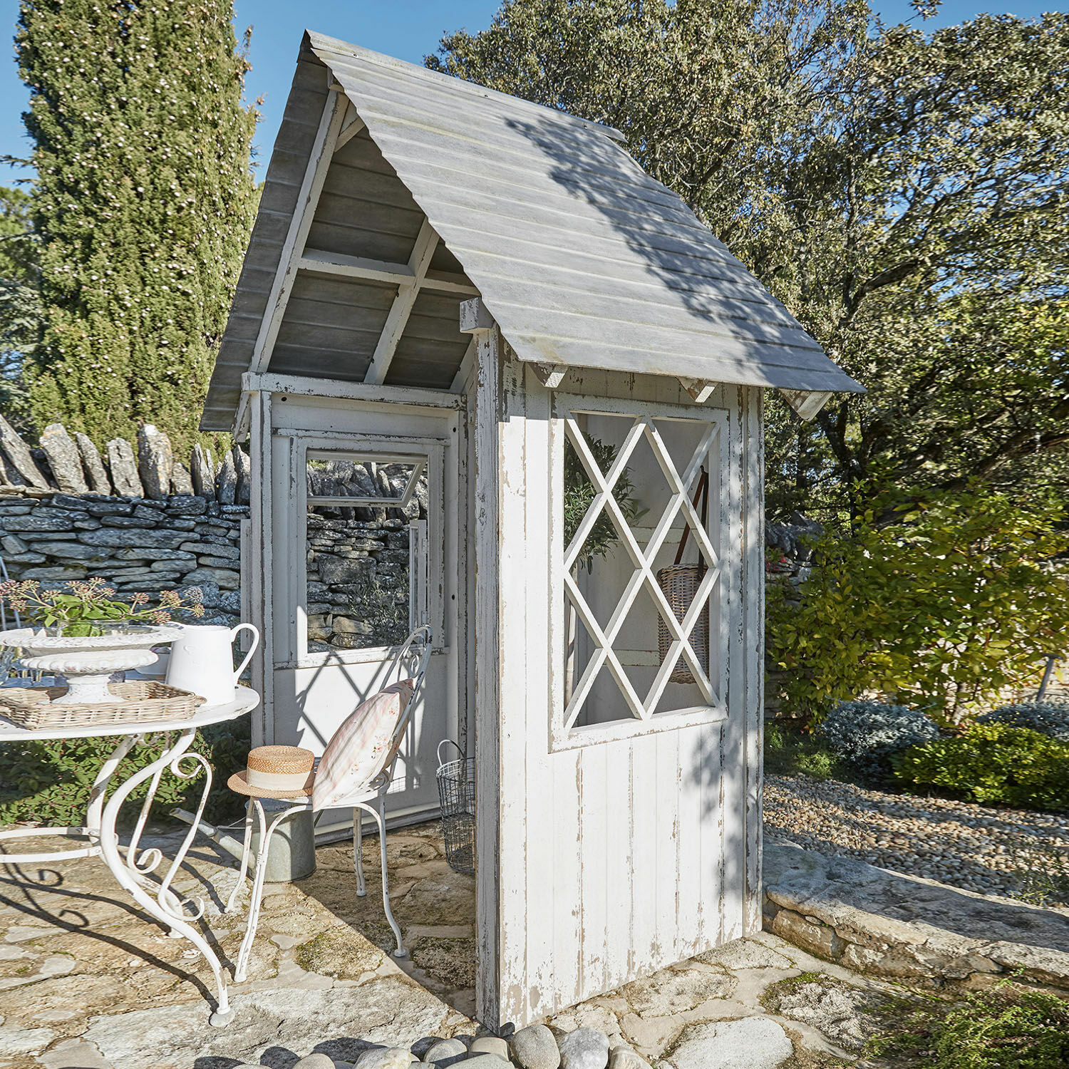 Weiße Holz-Gartenlaube mit Sprossenfenstern auf Steinboden in sonnigem Garten