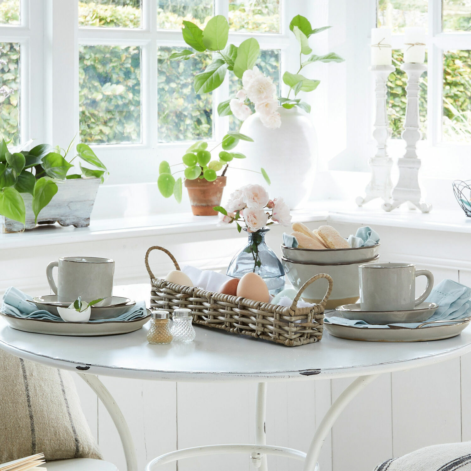 Helle Keramiktassen und Teller auf weißem Tisch mit Korb und Blumenarrangement als Dekor