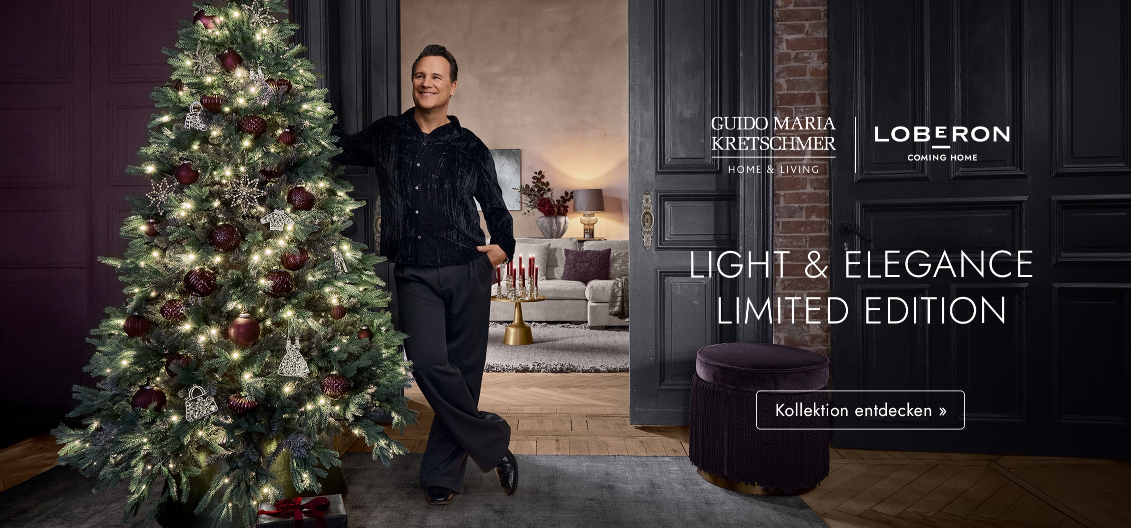 Mann lehnt an beleuchtetem Christbaum mit bordeauxroten und silbernen Kugeln in elegantem Wohnzimmer