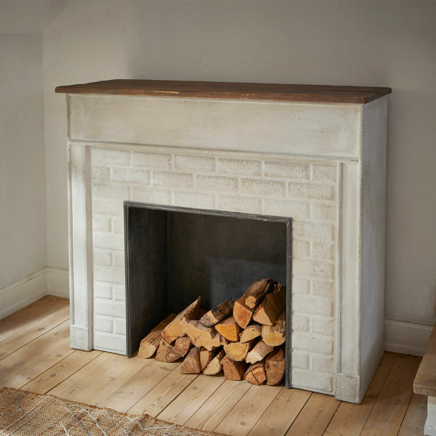 Weißer Beton-Kamin mit Holzplatte und gestapeltem Brennholz auf hellem Holzboden im rustikalen Stil